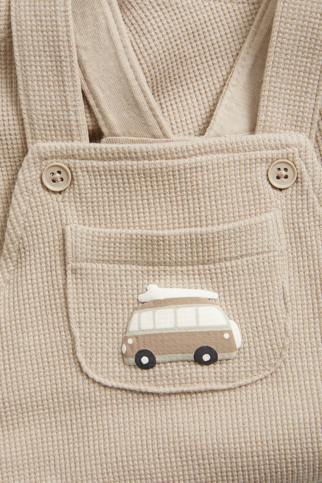Z przodu: beżowe waflowe ogrodniczki niemowlęce z guzikami i kieszenią z aplikacją kampera.