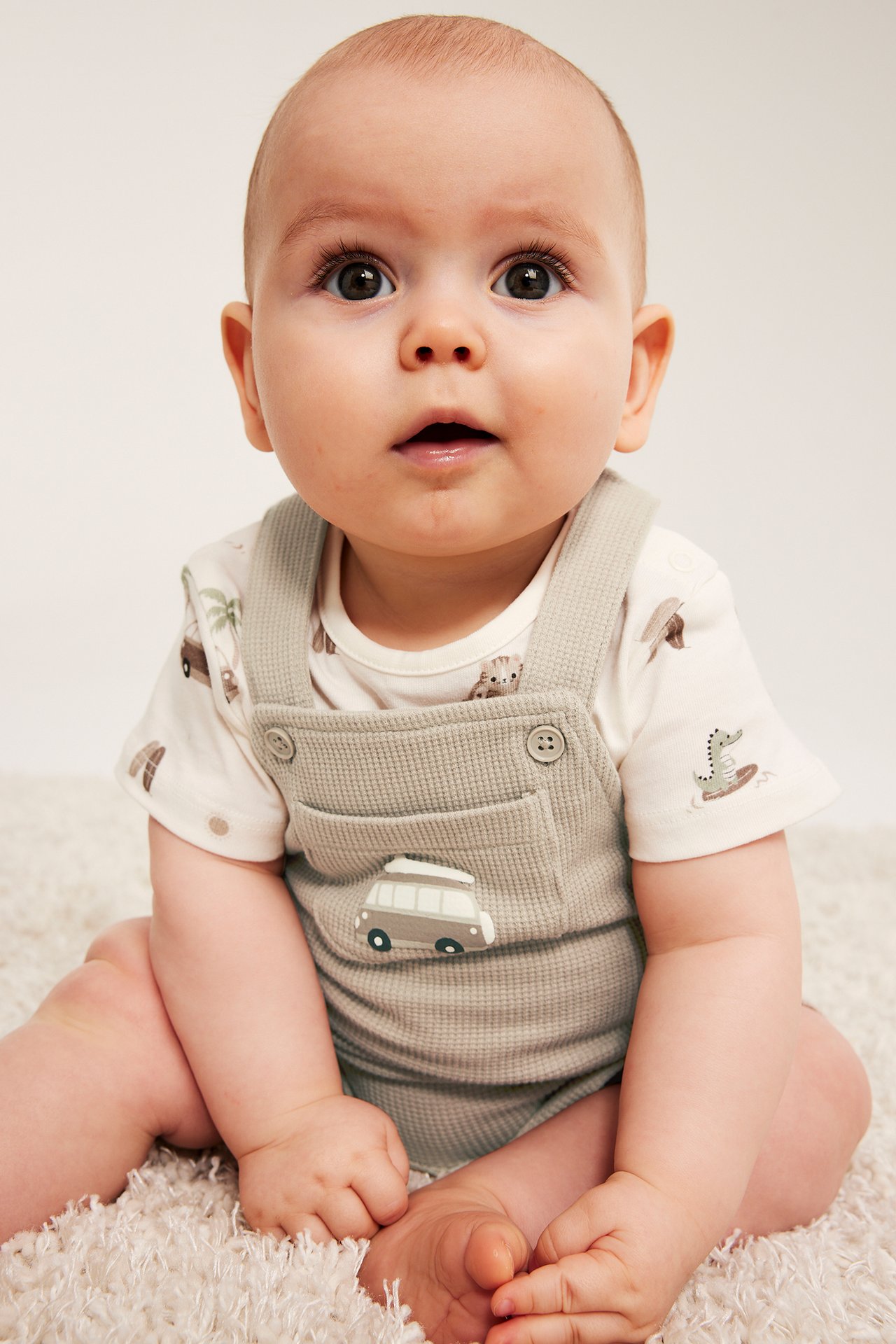 Przód: niemowlę w beżowych kombinezonach waflowych z kieszenią z autem, na wzorzystej koszulce.