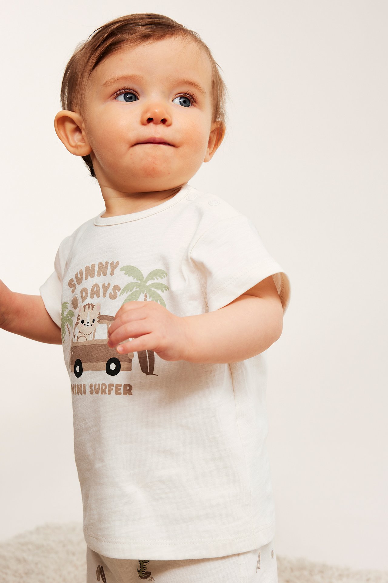 Baby w ecru dżersejowej koszulce z nadrukiem 'Sunny Days', zatrzaskami na ramieniu i szortach.