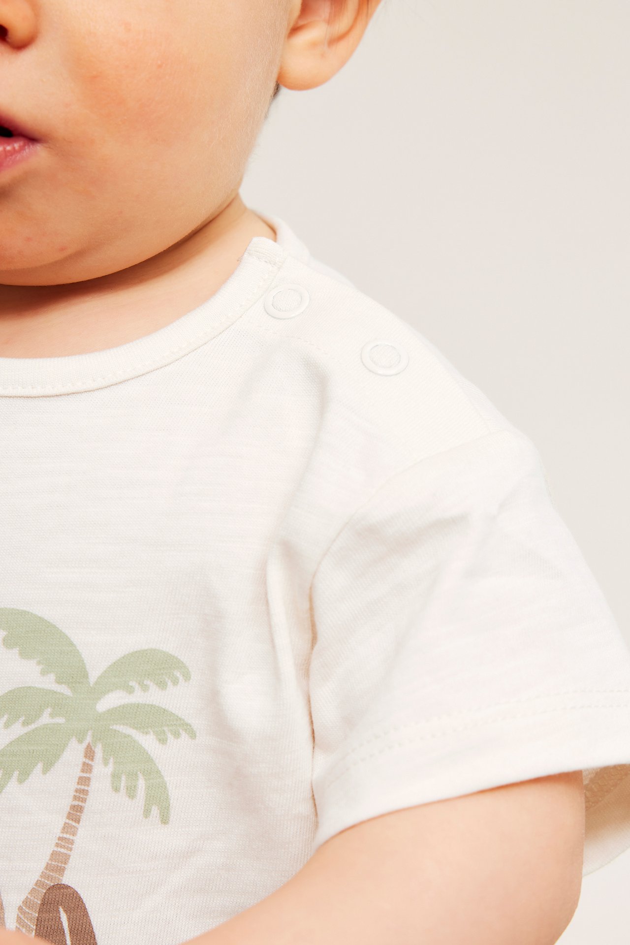Närbild av baby i offwhite bomulls-t-shirt med palmtryck och axelknappar.