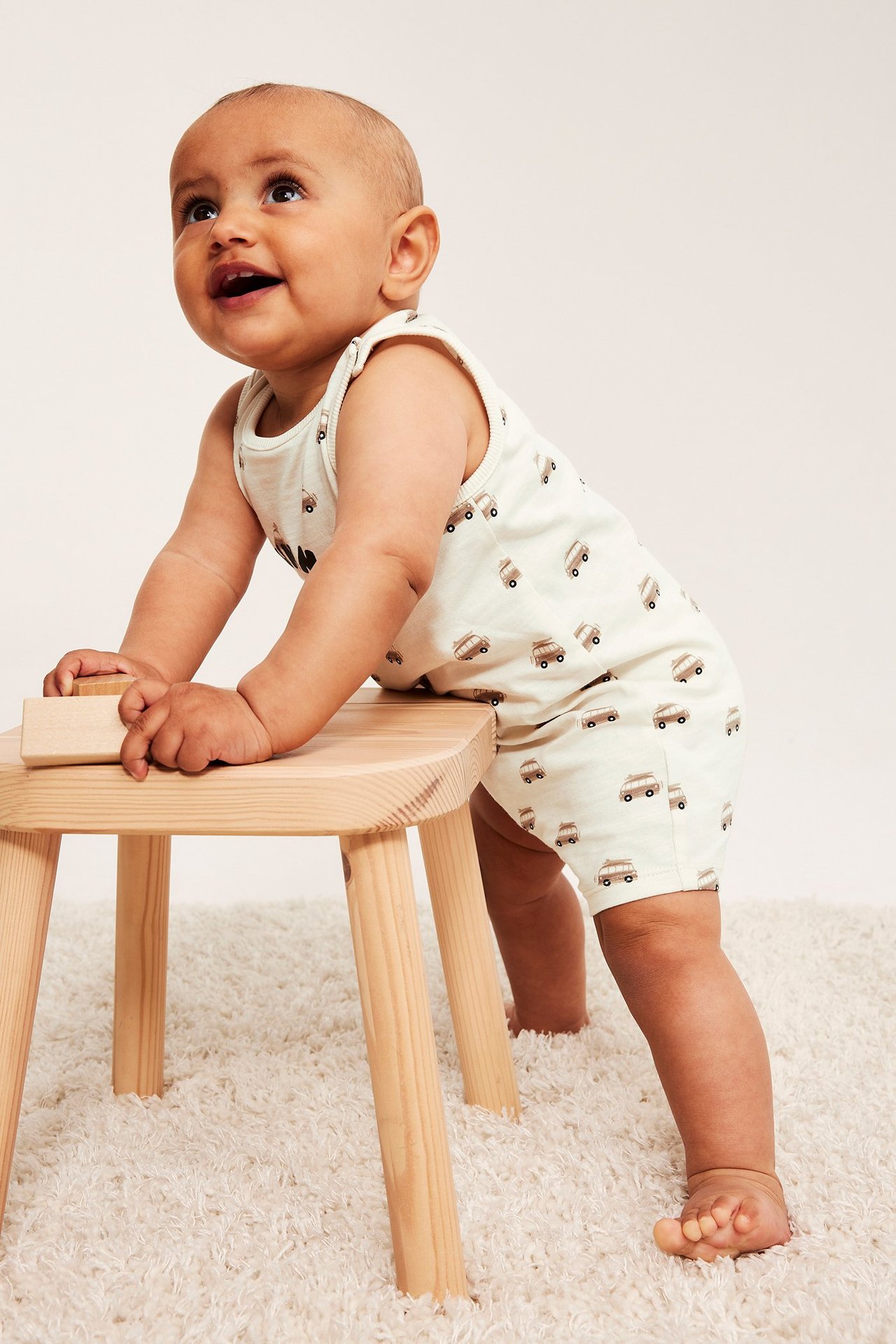 Baby i offwhite bomulls-jumpsuit med bussmönster, håller träklossar på en ljus träpall.