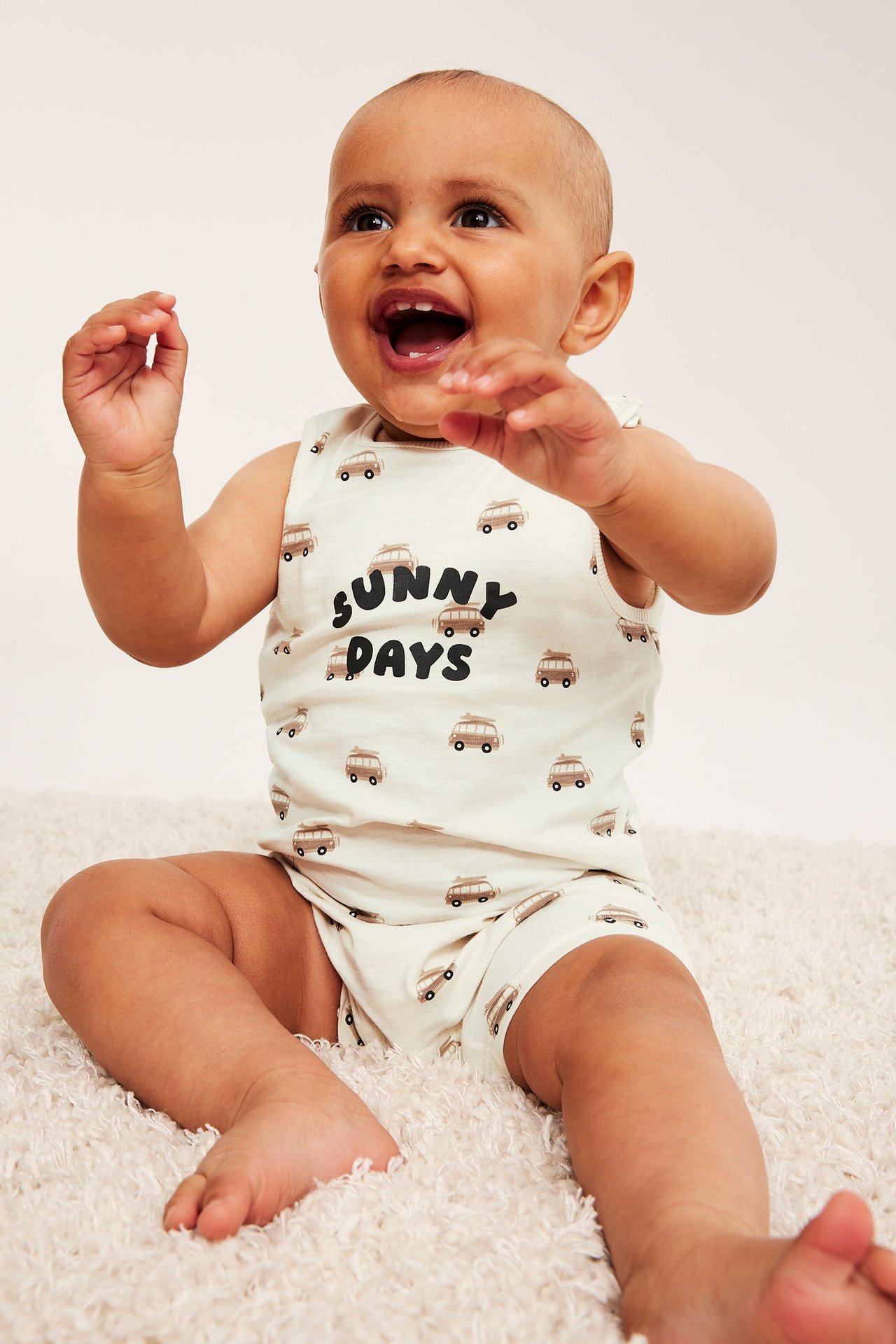 Leende baby i en benvit ärmlös bomullsjumpsuit med brunt bussmönster och texten 'Sunny Days'.