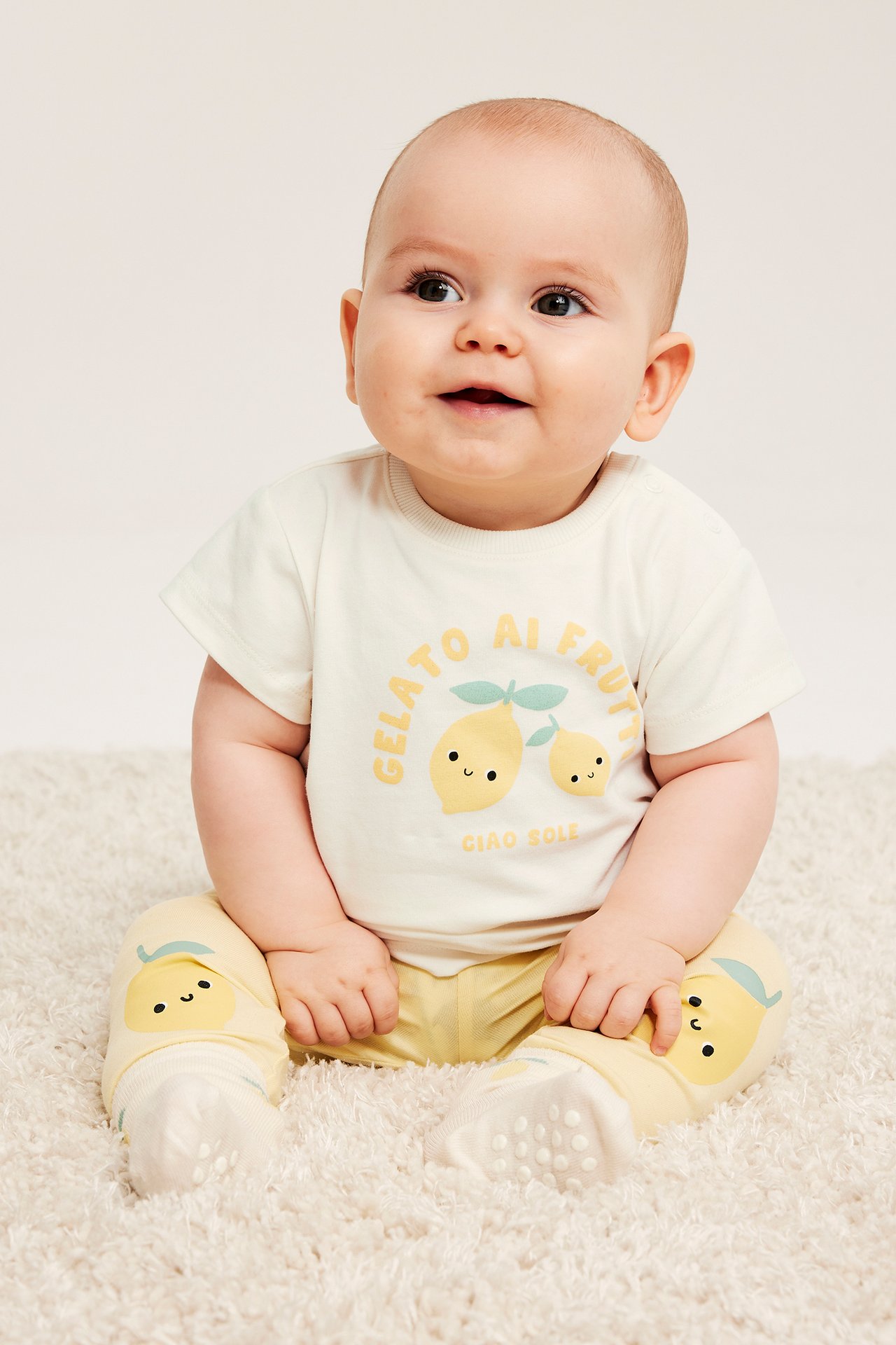 Framsida av baby i en offwhite t-shirt med citrontryck och tryckknappar, samt gula leggings.