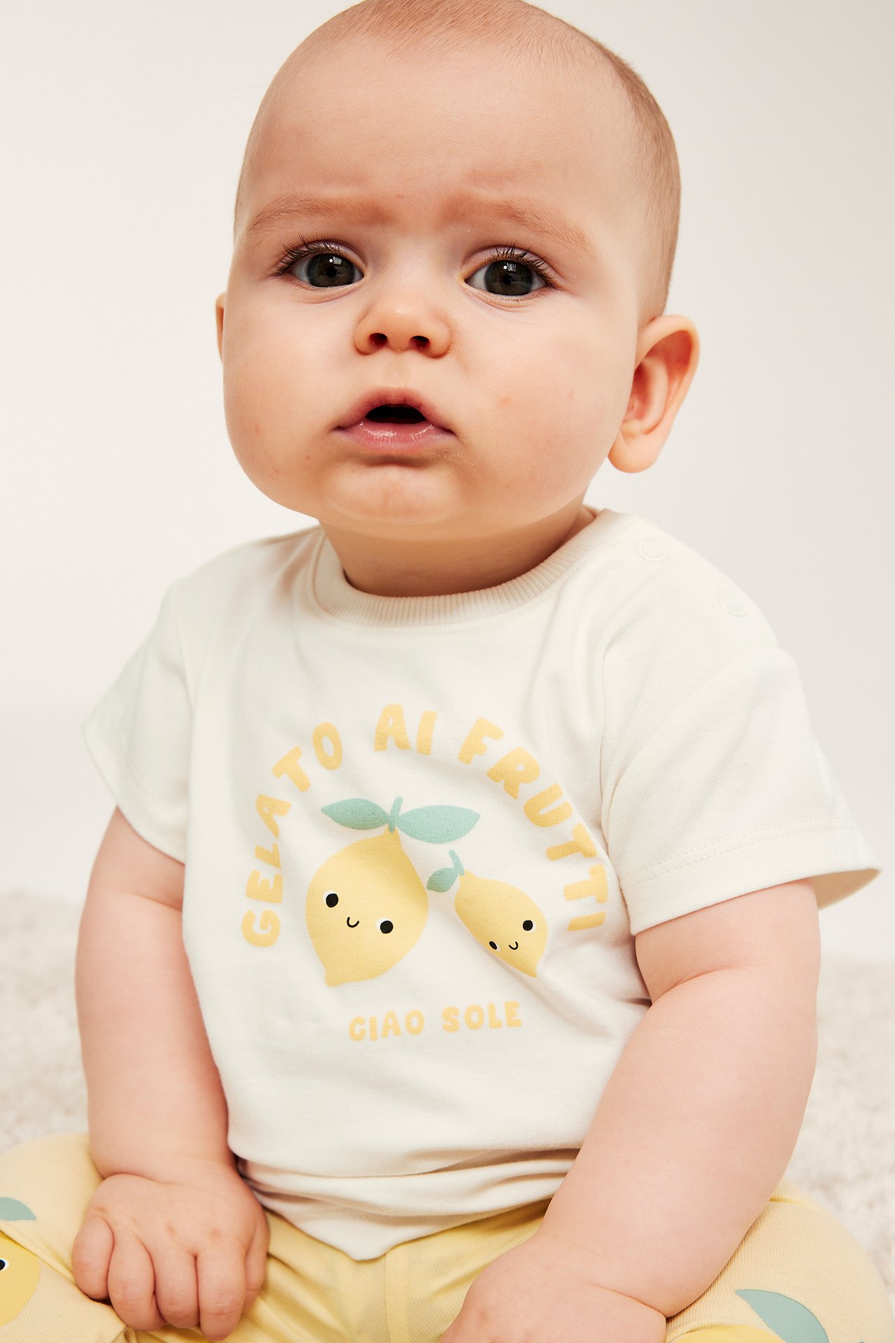 Frontbilde av en baby i en offwhite t-skjorte med sitrontrykk, matchet med gule sitronmønstrede bukser.