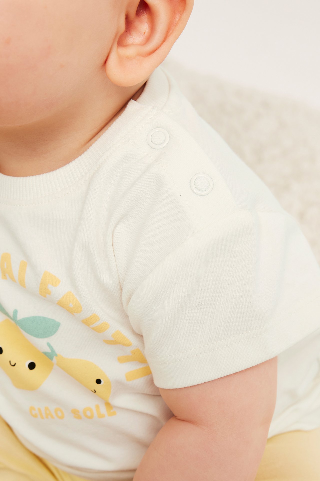 Närbild av baby i en offwhite t-shirt i bomullsblandning med frukttryck och tryckknappar på axeln.