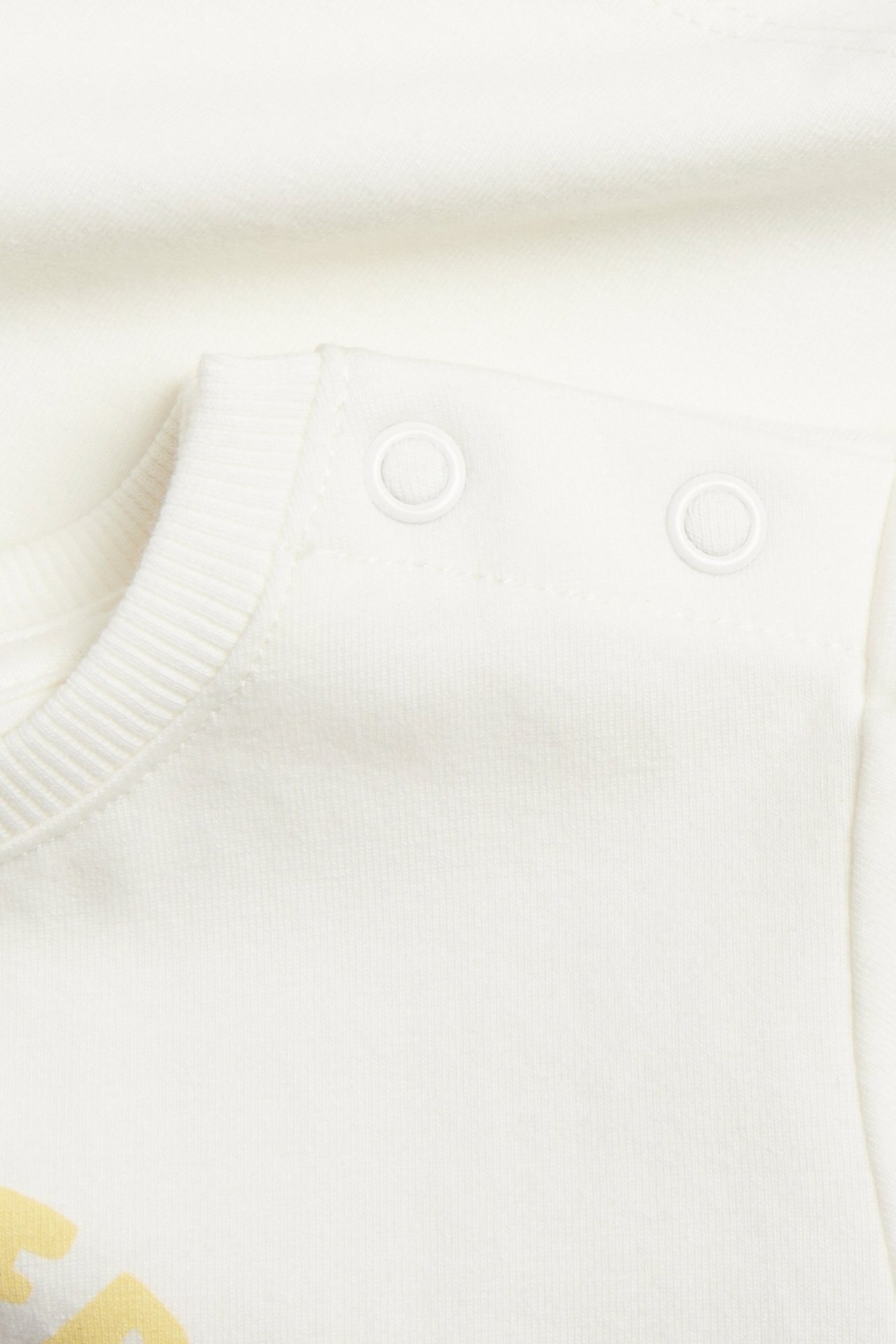Närbild av en offwhite kortärmad babyt-shirt med tryckknappar på axeln och gult tryck fram.