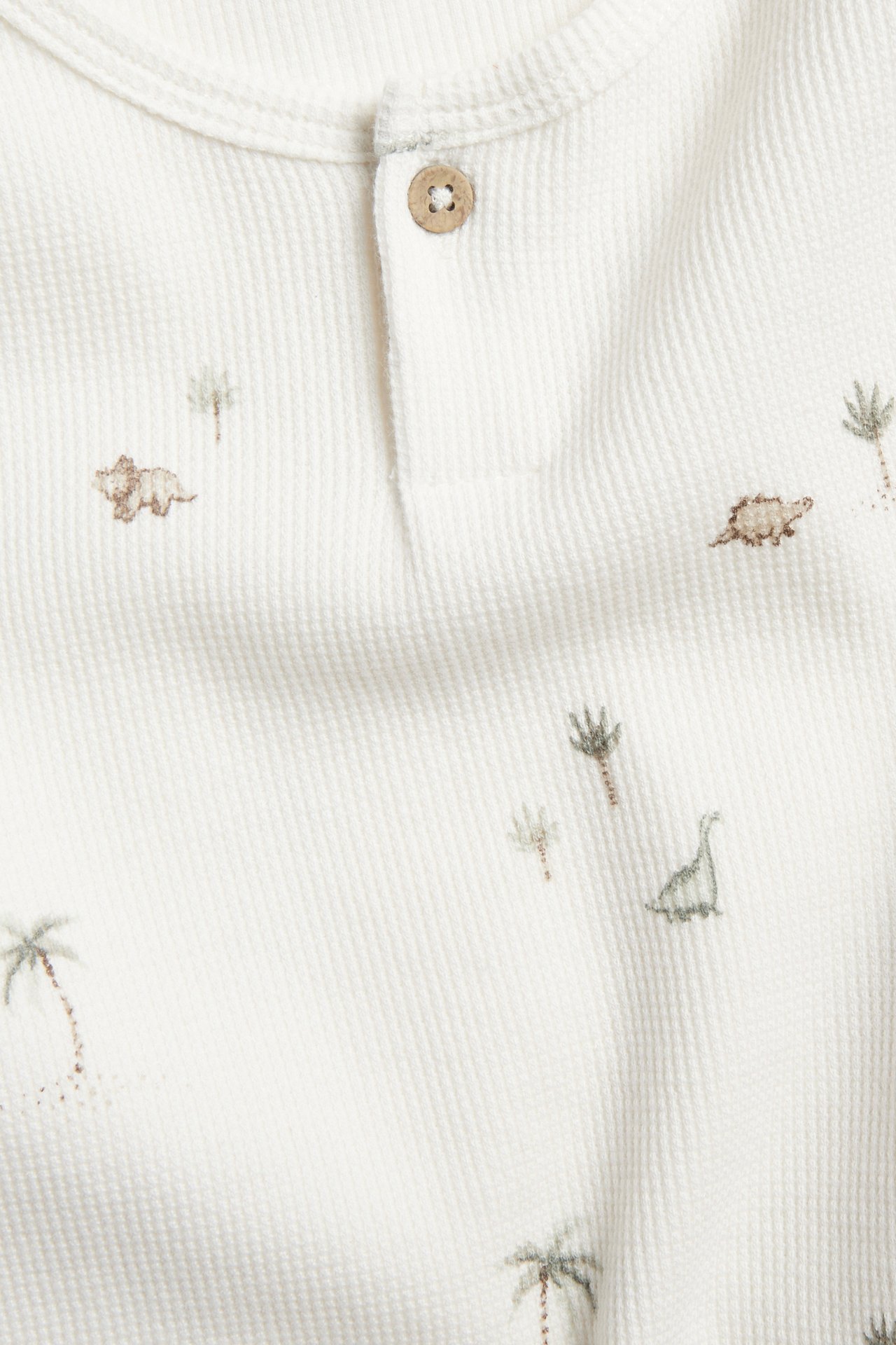 Närbild: Offwhite våfflad långärmad t-shirt med knappslå, dinosaurie- och palmträdsmönster.