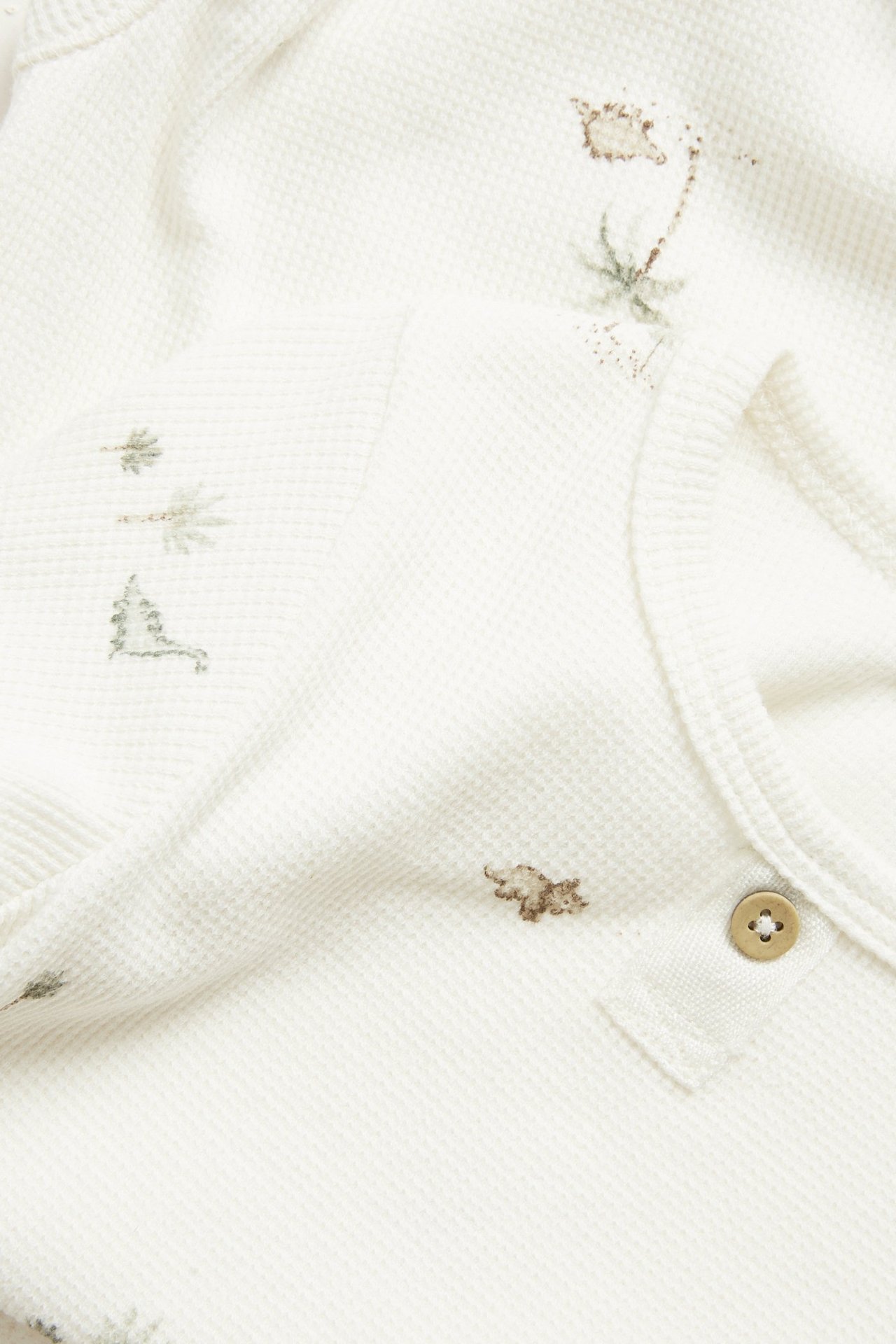 Lähikuva luonnonvalkoisesta vauvan vohvelibodysta, brodeerattuja dinosauruksia, palmuja ja koristenappi.