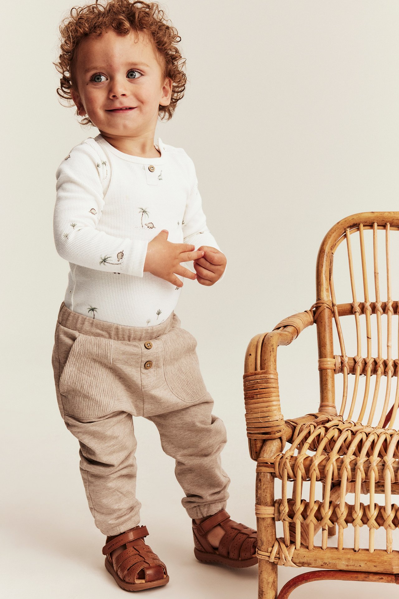 Frontutsikt, baby i offwhite vaffelstrikket langermet body med dinosaurmønster, beige ribbestrikkede bukser og brune skinnsandaler.