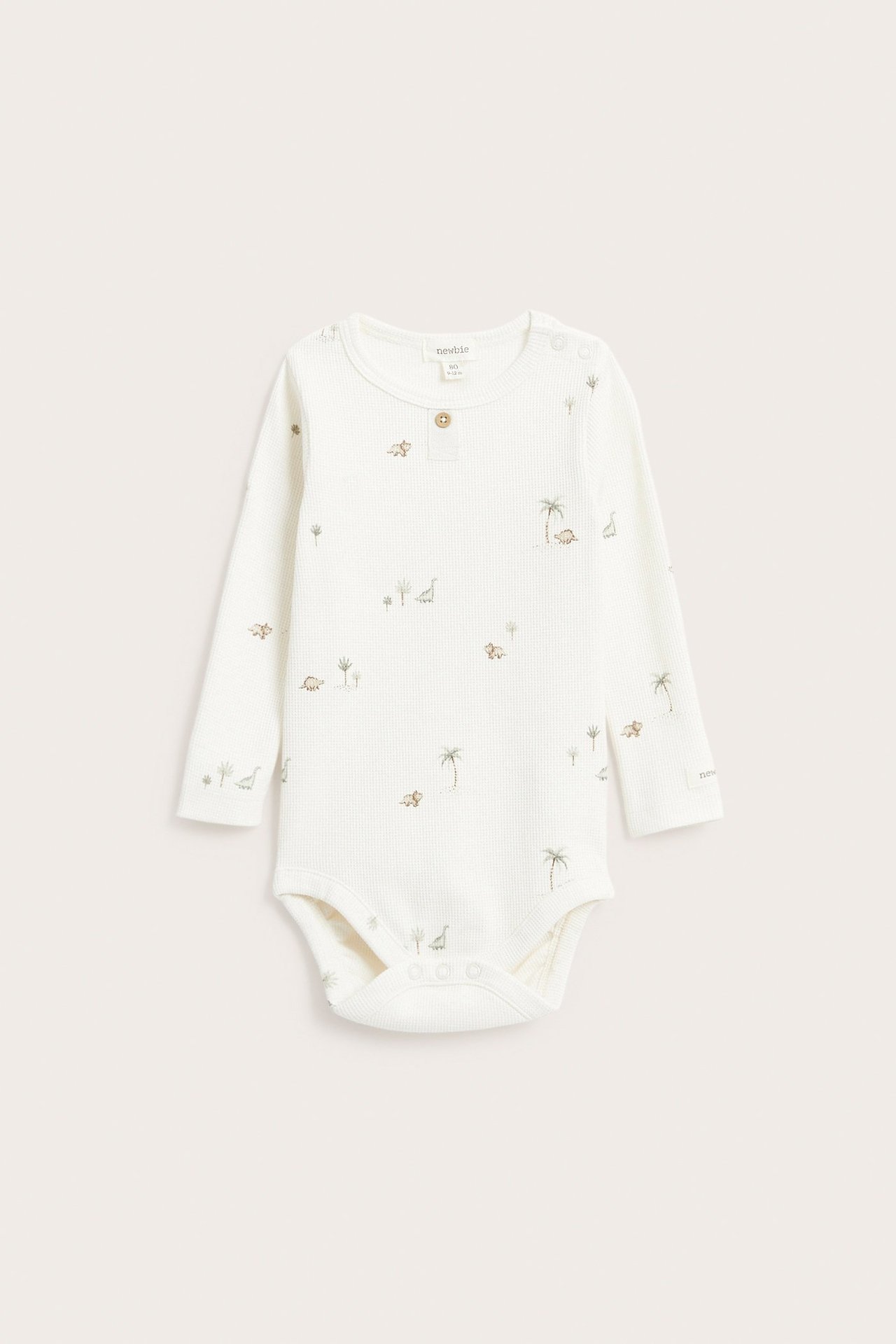 Edestä näkymä luonnonvalkoisesta pitkähihaisesta vohvelineulotusta vauvanbodysta dinosaurus- ja palmukuvioilla.