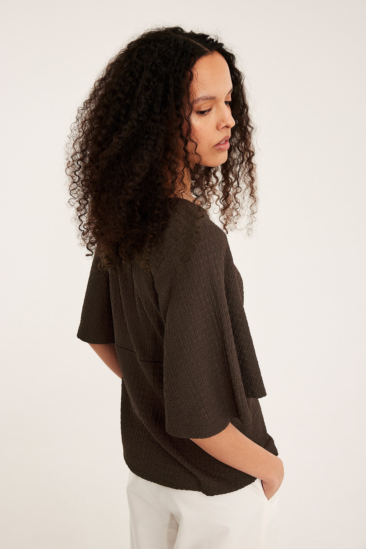 Sideutsikt av en kvinne i mørk brun strukturert kortermet topp med vide ermer, kombinert med hvite bukser.