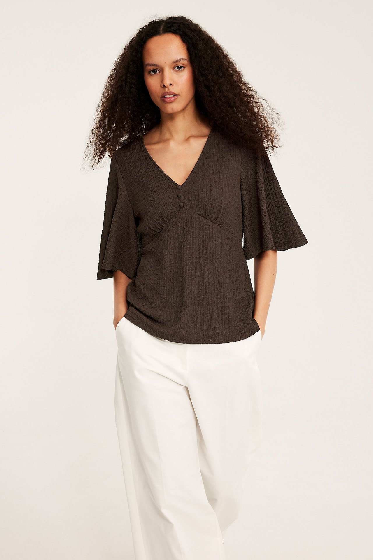Framifrån: kvinna i mörkbrun strukturerad V-ringad topp med vida ärmar och vita byxor.