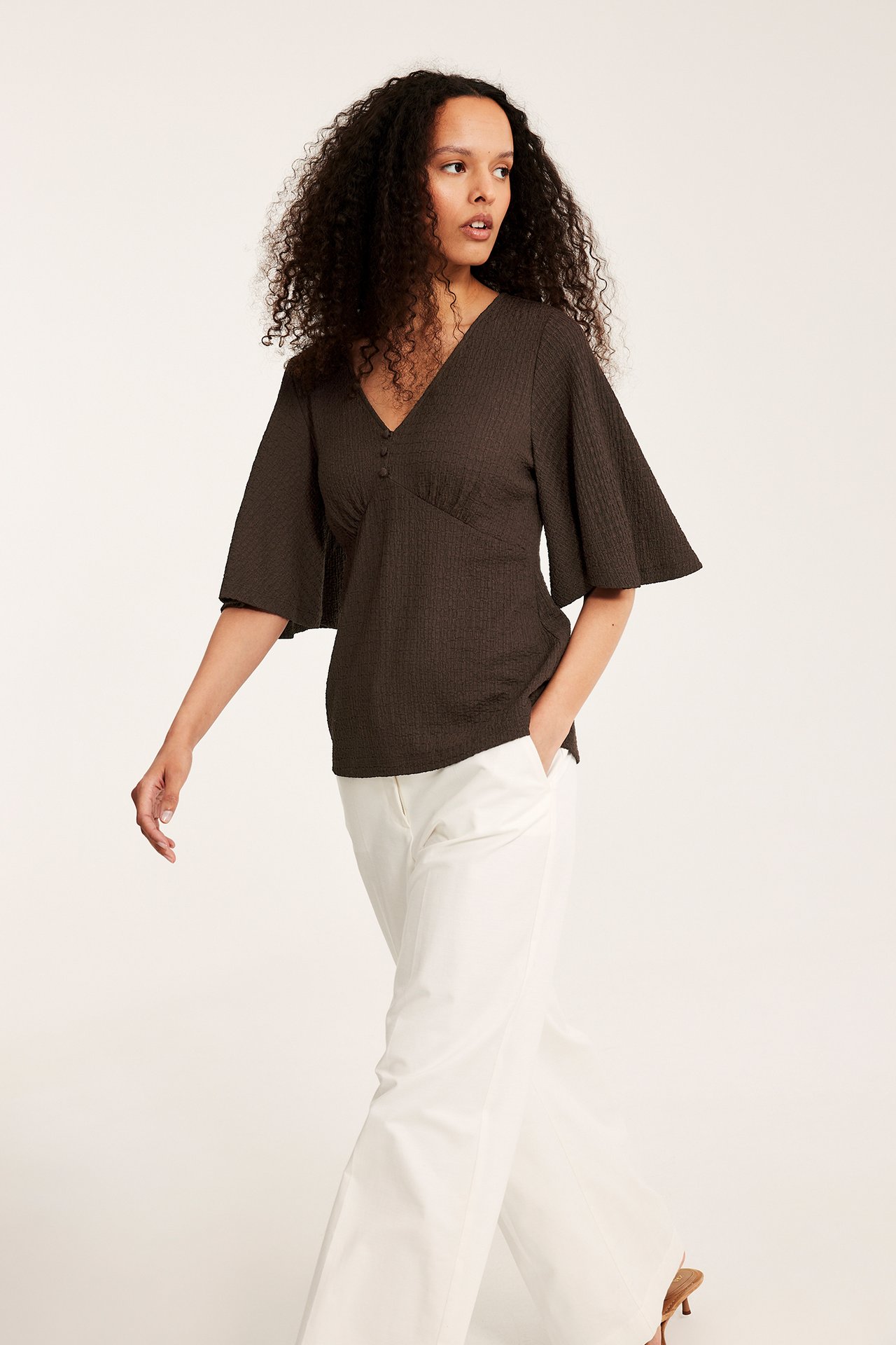 Framifrån: kvinna i mörkbrun strukturerad V-ringad topp med vida ärmar och vita byxor med vida ben.