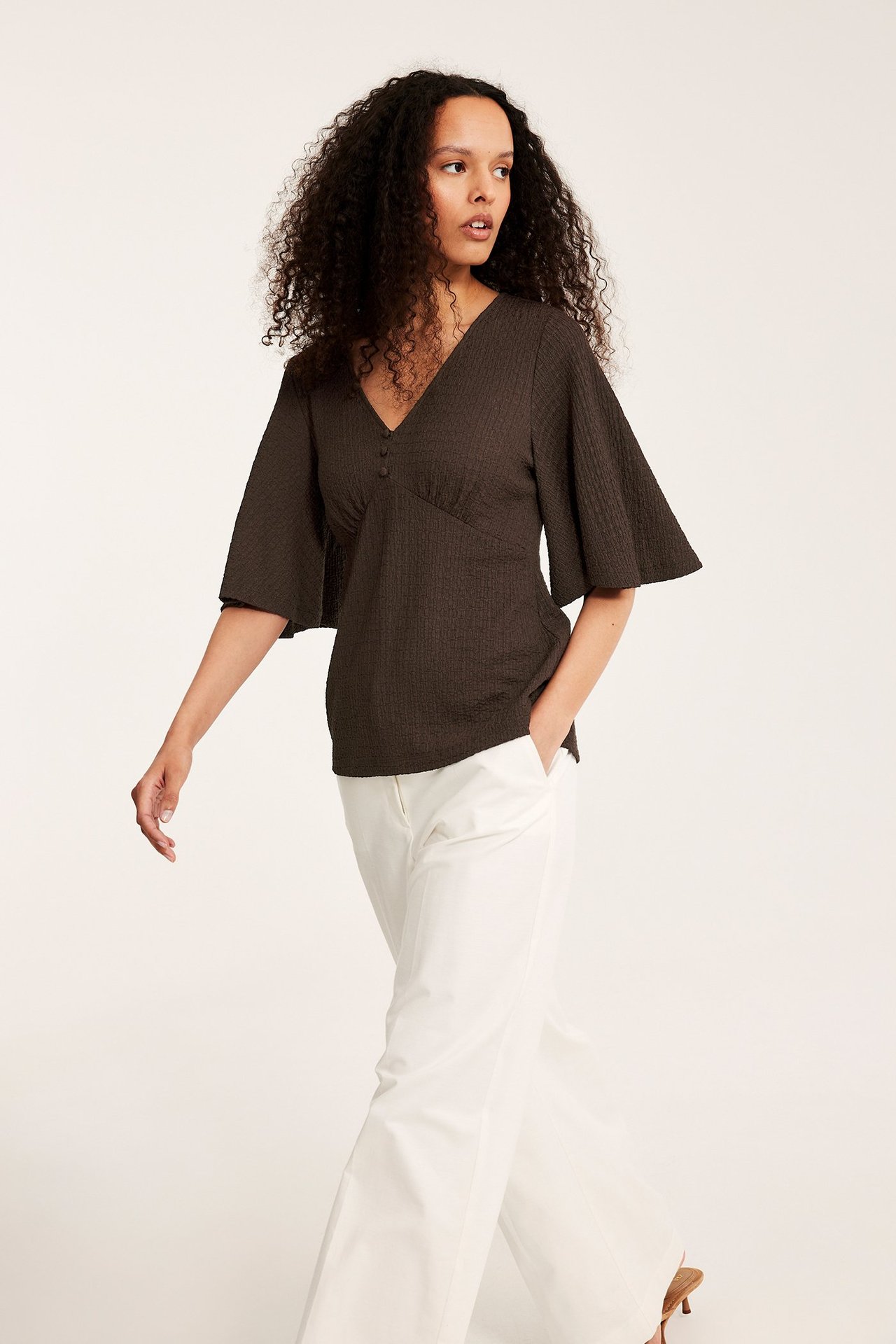 Framifrån: kvinna i mörkbrun strukturerad V-ringad topp med vida ärmar och vita byxor med vida ben.