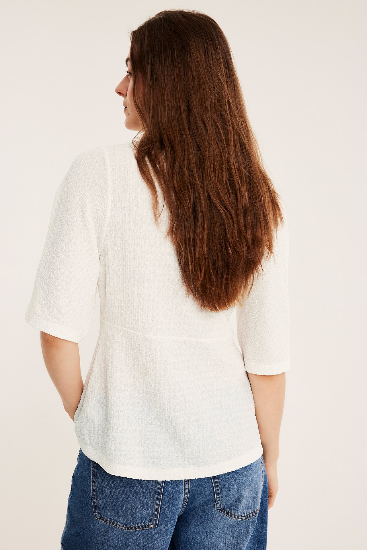 Naisen selkäpuolen näkymä yllään luonnonvalkoinen strukturoitu lyhythihainen toppi ja siniset farkut.