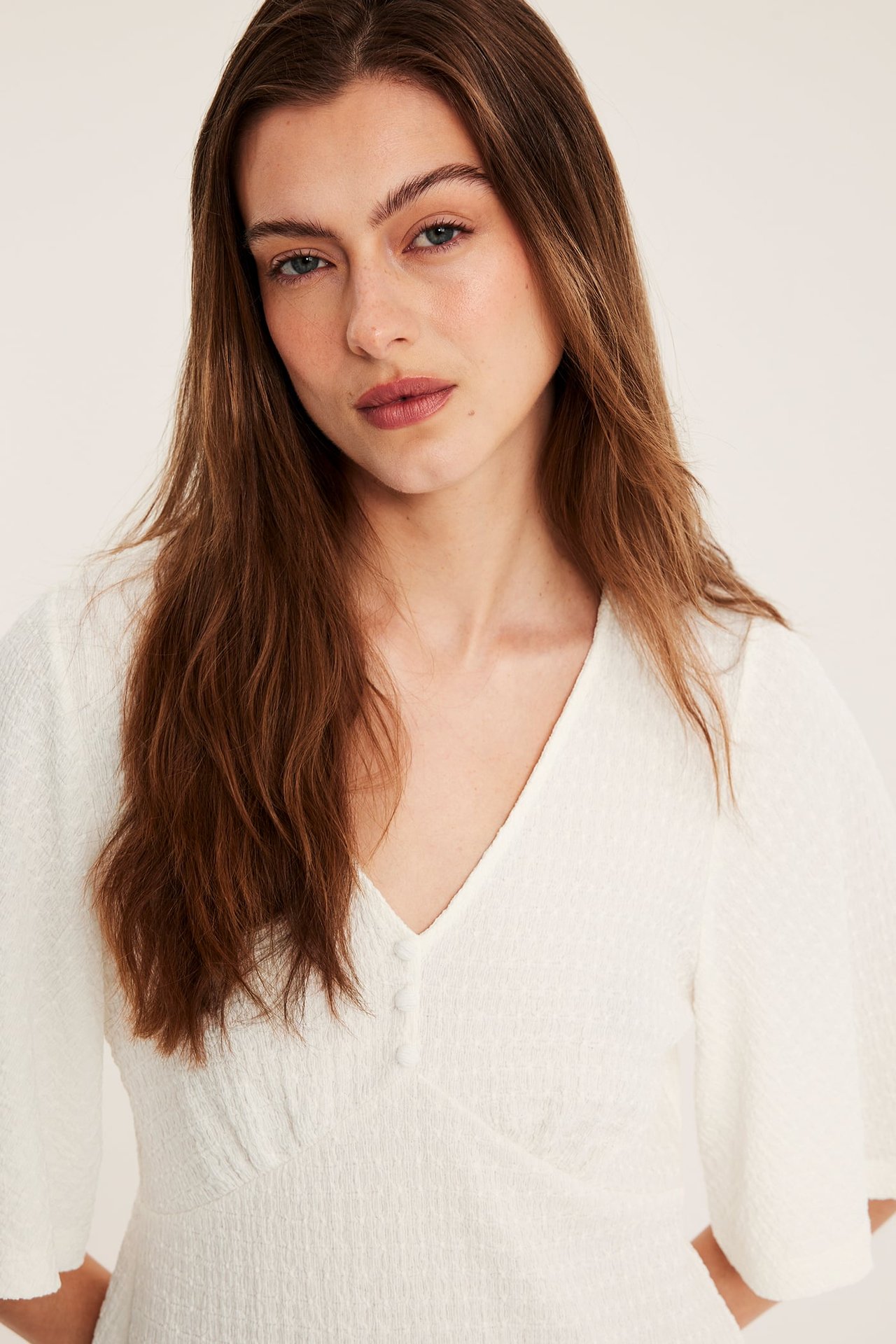 Närbild framifrån på kvinna i offwhite strukturerad V-ringad topp med vida ärmar och knappar fram.