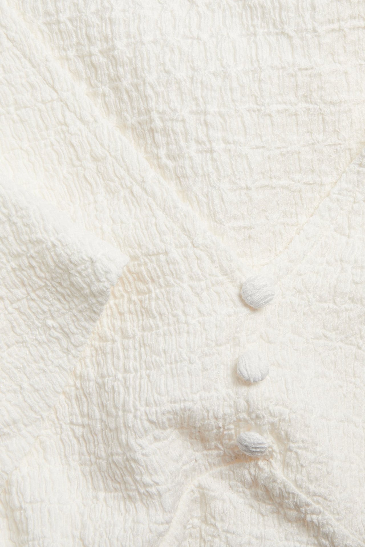 Zbliżenie na kremowy, strukturalny top z dekoltem w serek i trzema guzikami pokrytymi tkaniną.