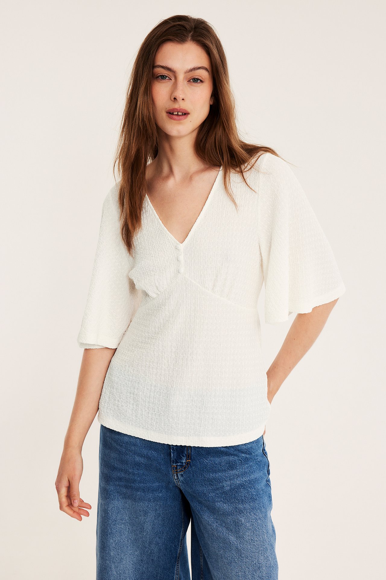Naisen etunäkymä: luonnonvalkoinen strukturoitu V-kaula-aukkoine toppi leveillä hihoilla ja siniset farkut.