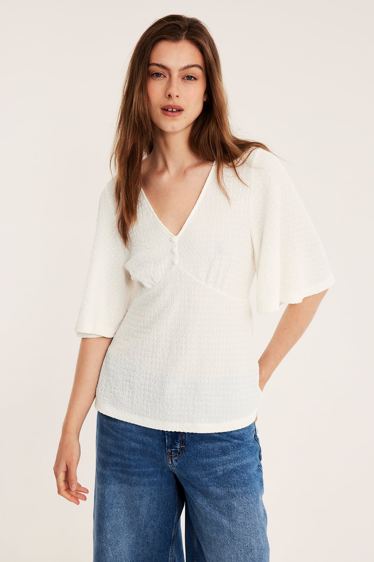 Frontvy av en kvinna i offwhite strukturerad V-ringad topp med vida ärmar och blå jeans.
