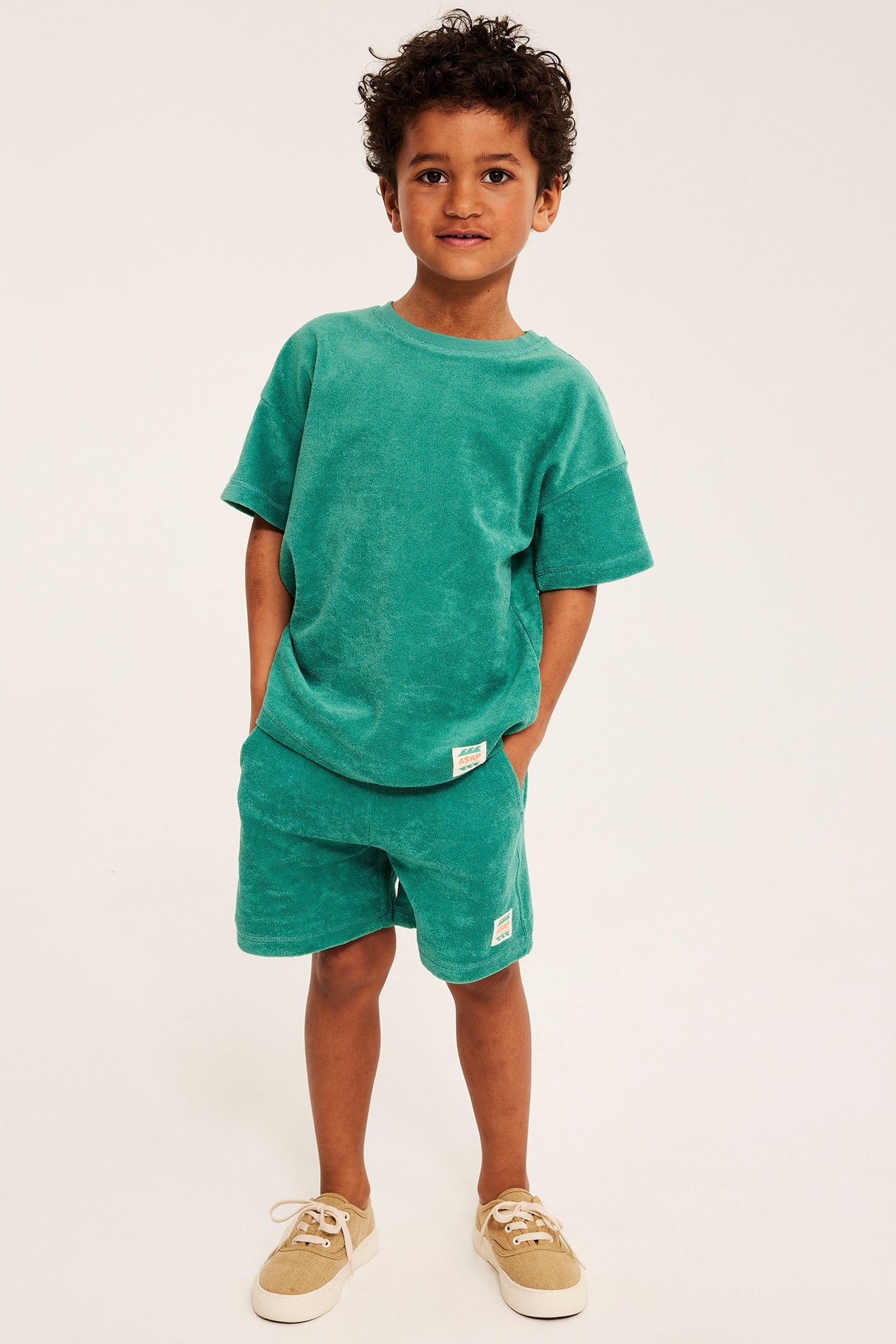 Framifrån: barn i en oversize grön frotté-t-shirt, matchande shorts och beige sneakers.