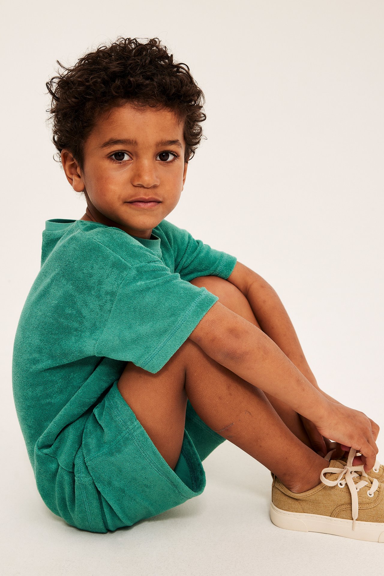Widok z przodu: dziecko w zielonym, luźnym T-shircie frotte, pasujących szortach i beżowych trampkach.
