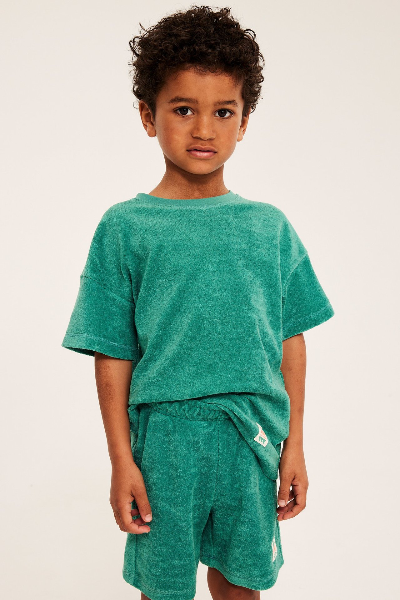 Frontvy av ett barn i grön oversized frotté-t-shirt och matchande gröna frottéshorts.