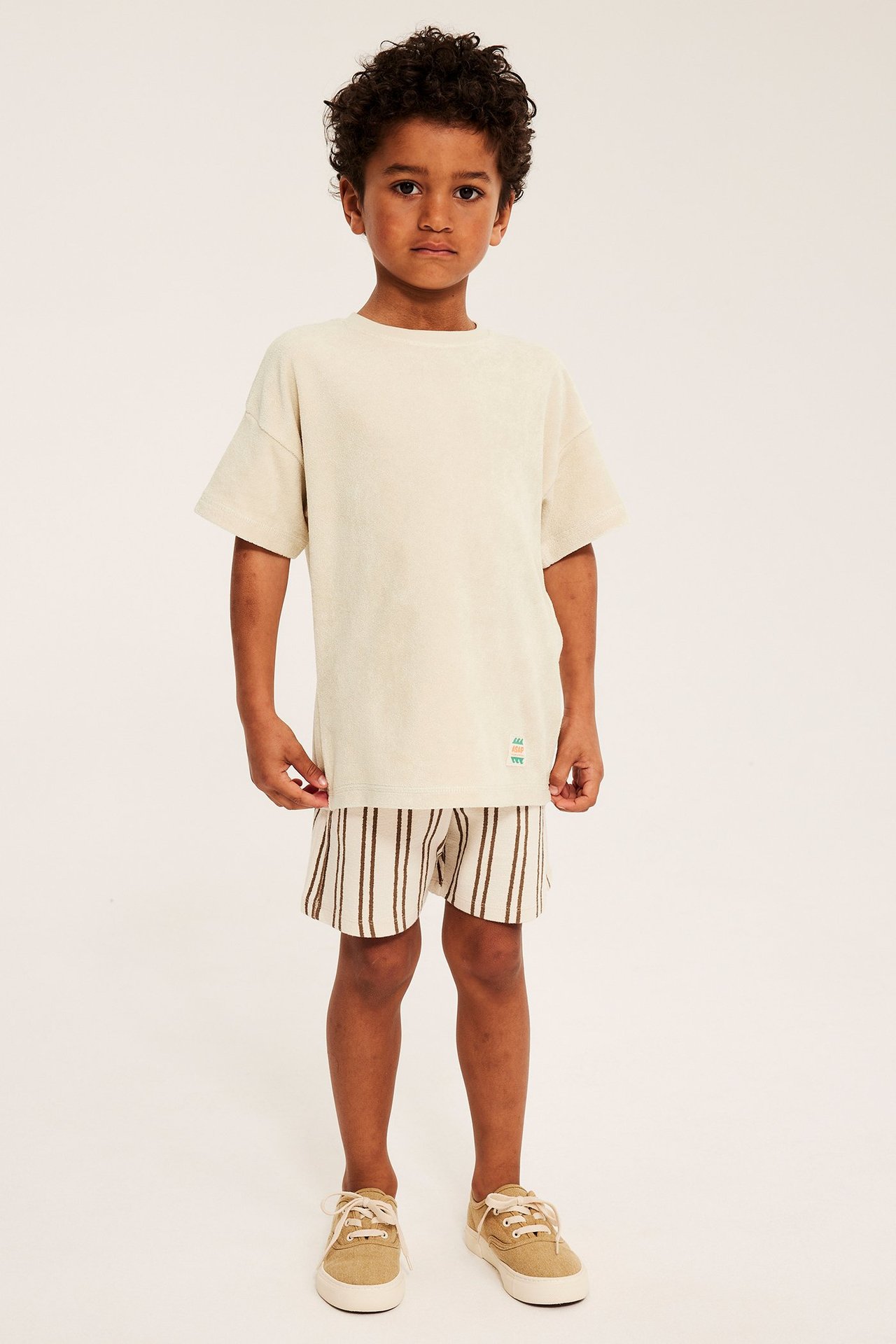 Framsida: Ett barn i ljusbeige oversize frotté t-shirt med patch, randiga shorts och beiga sneakers.