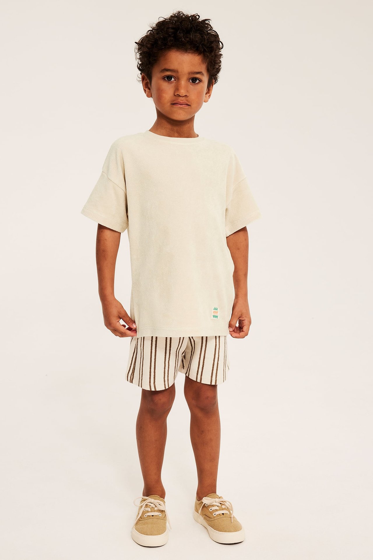 Framsida: Ett barn i ljusbeige oversize frotté t-shirt med patch, randiga shorts och beiga sneakers.