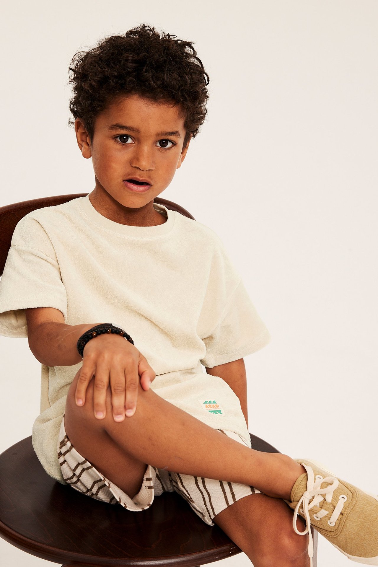 Forfra: et barn i lys beige oversized frotté-t-skjorte, stripete shorts, svart perlearmbånd og beige sneakers.