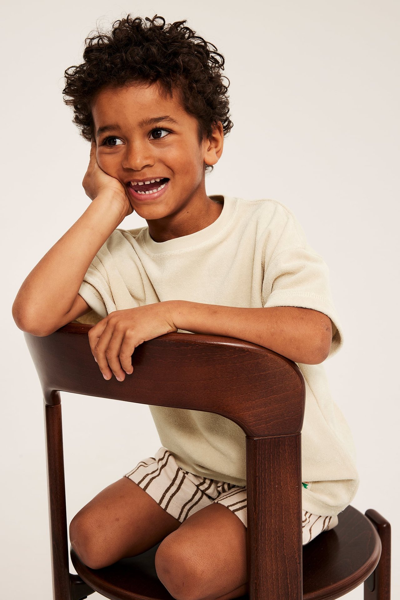 Framifrån: barn i ljusbeige frotté T-shirt och randiga shorts, sittande på en stol.