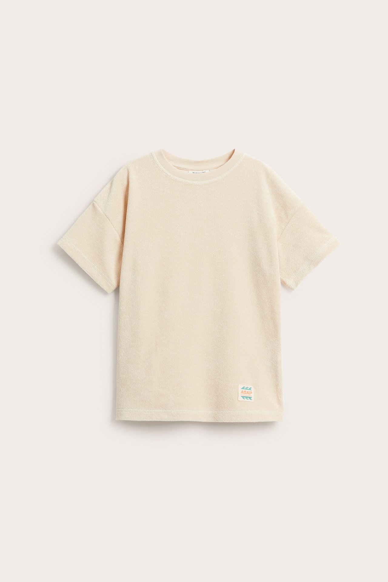 Framifrån en ljusbeige kortärmad frotté t-shirt med oversize passform, droppade axlar och liten patch.