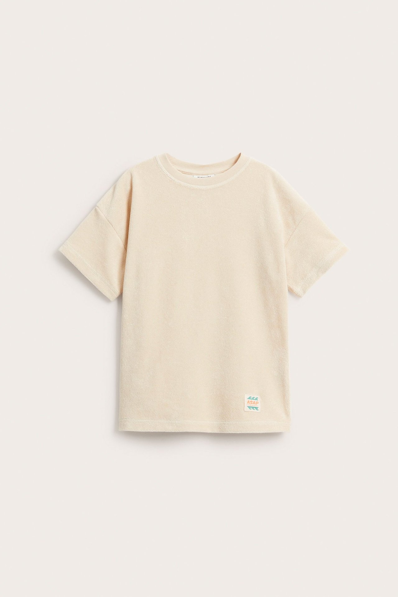 Widok z przodu jasnobeżowej koszulki frotte z krótkim rękawem, luźnym fasonem, obniżonymi ramionami i naszywką.