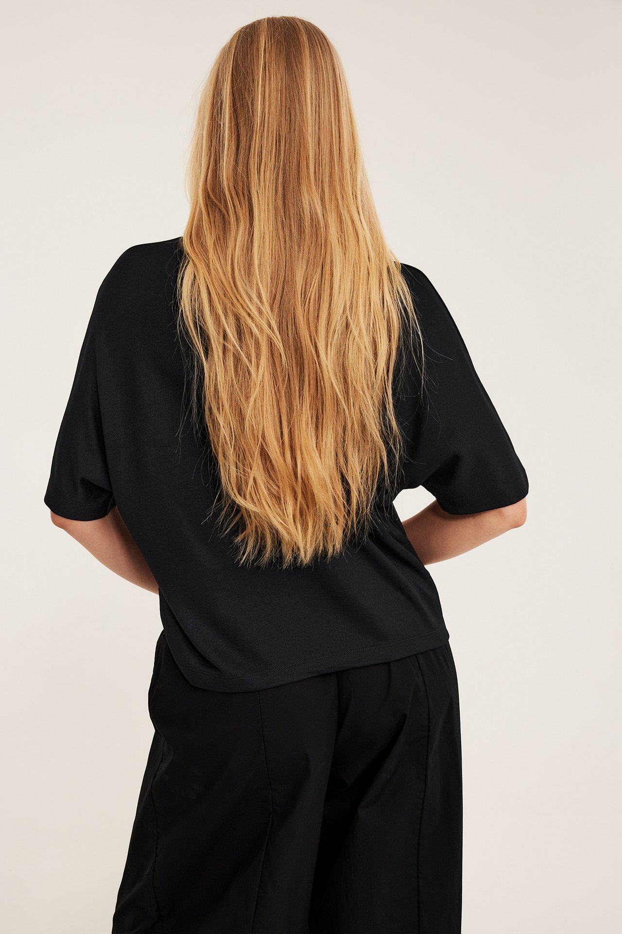 Nainen takaa, yllään oversized musta viskoosisekoite lyhythihainen toppi ja mustat leveälahkeiset housut.