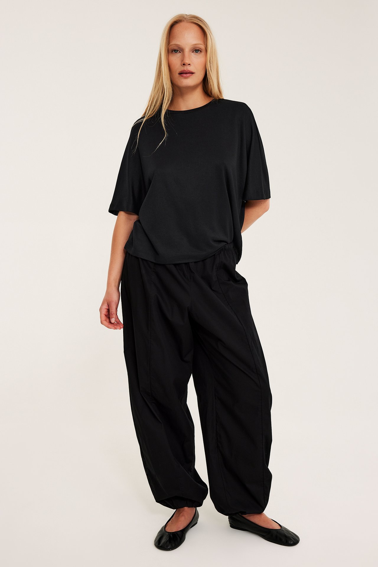 Naisen etukuva, yllään musta oversize-toppi lyhyillä hihoilla, mustat tilavat housut ja mustat ballerinakengät.