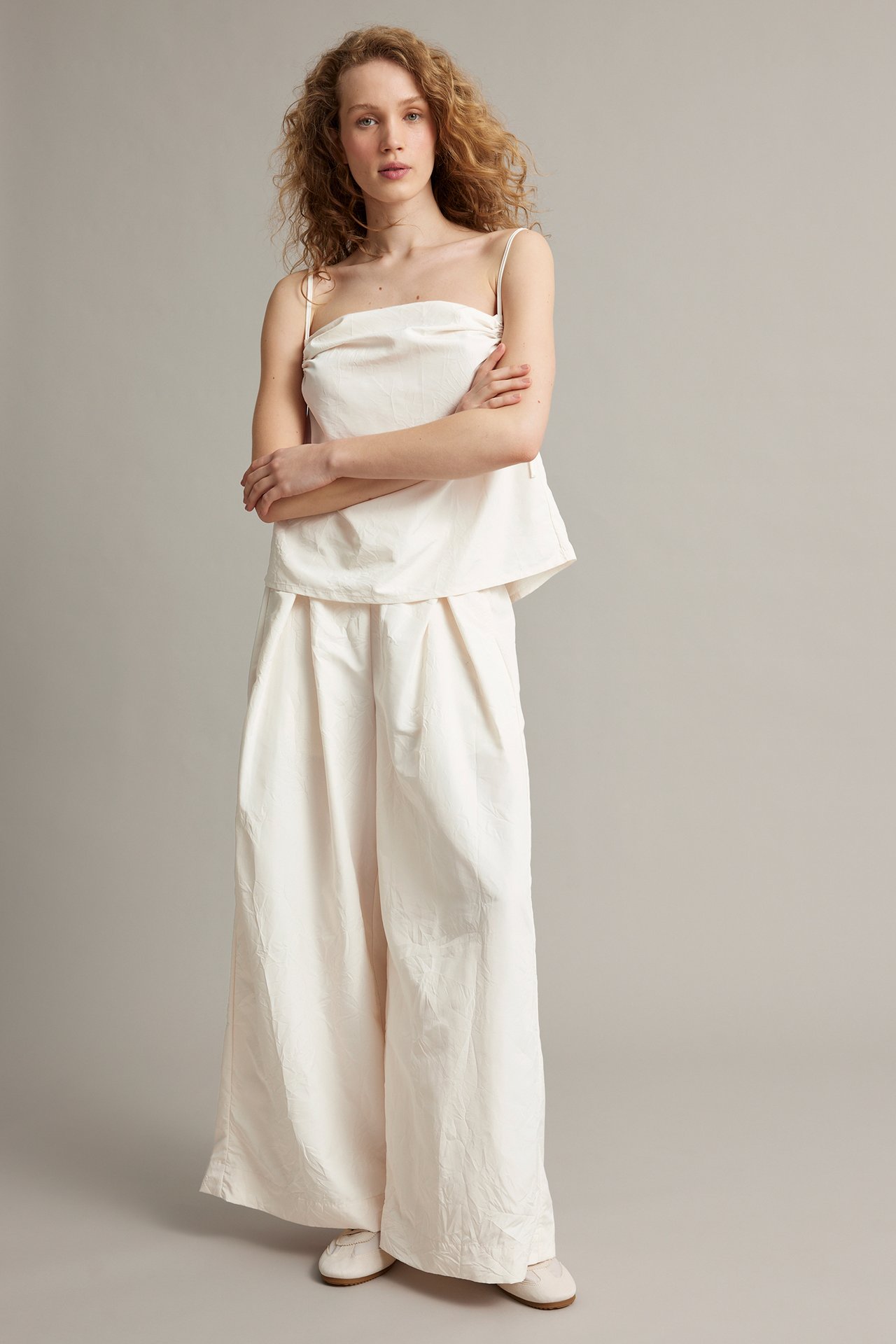 Frontvy: Kvinna i vita vida polyesterbyxor med motveck, vit camisole och ljusa platta skor.