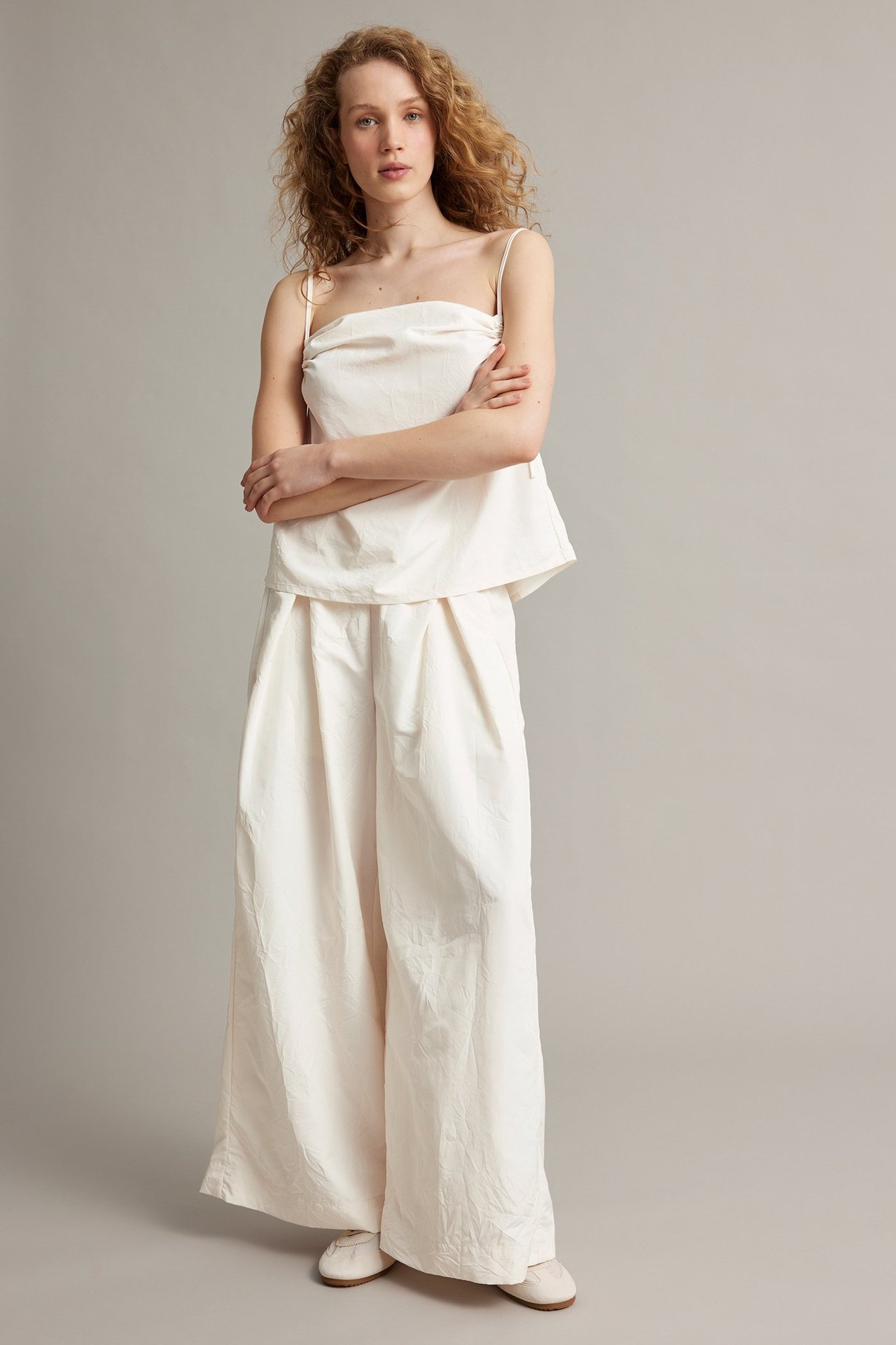 Frontvy: Kvinna i vita vida polyesterbyxor med motveck, vit camisole och ljusa platta skor.