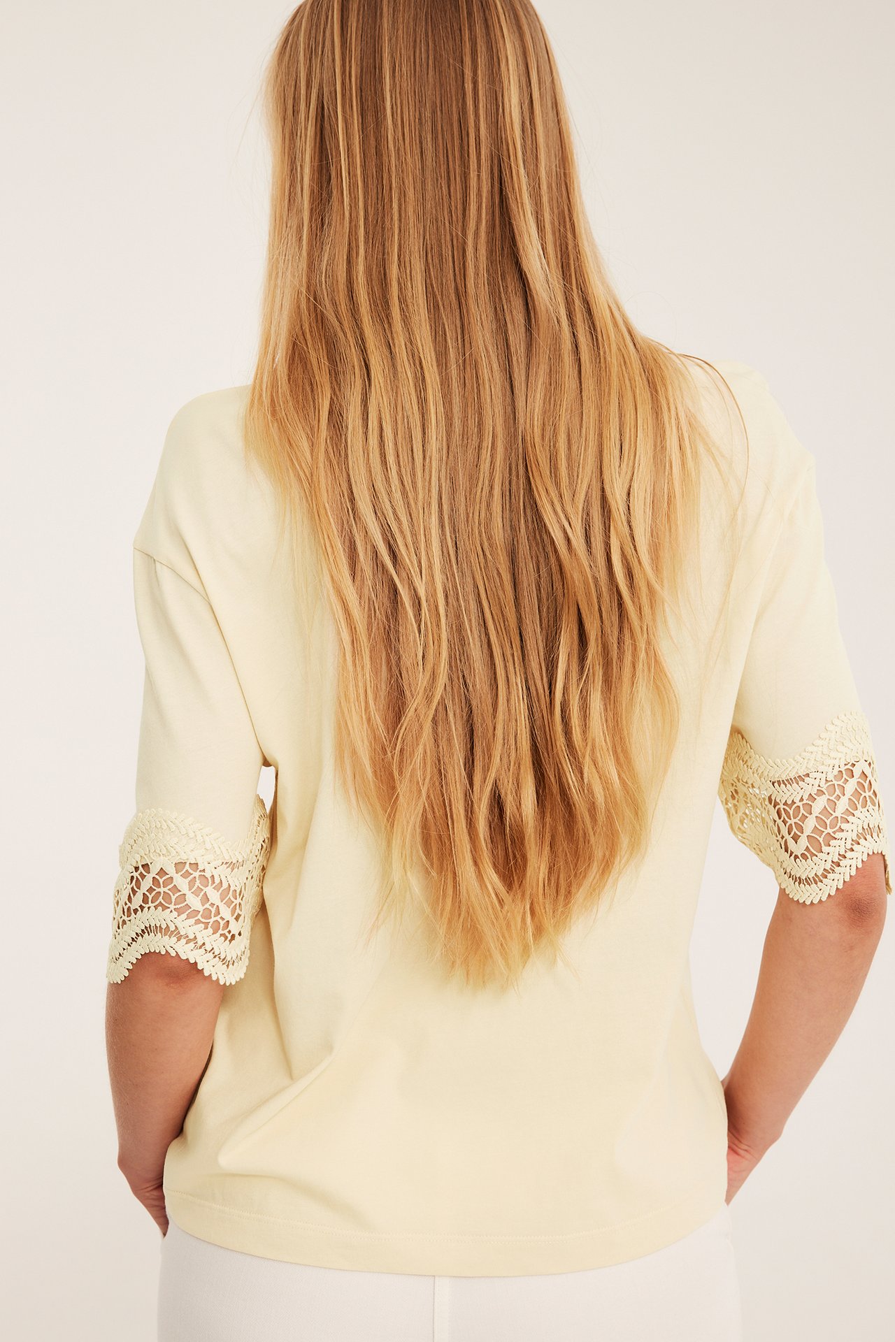 Bakfra: kvinne i lys gul løs t-skjorte med blondearmer og hvite bukser.