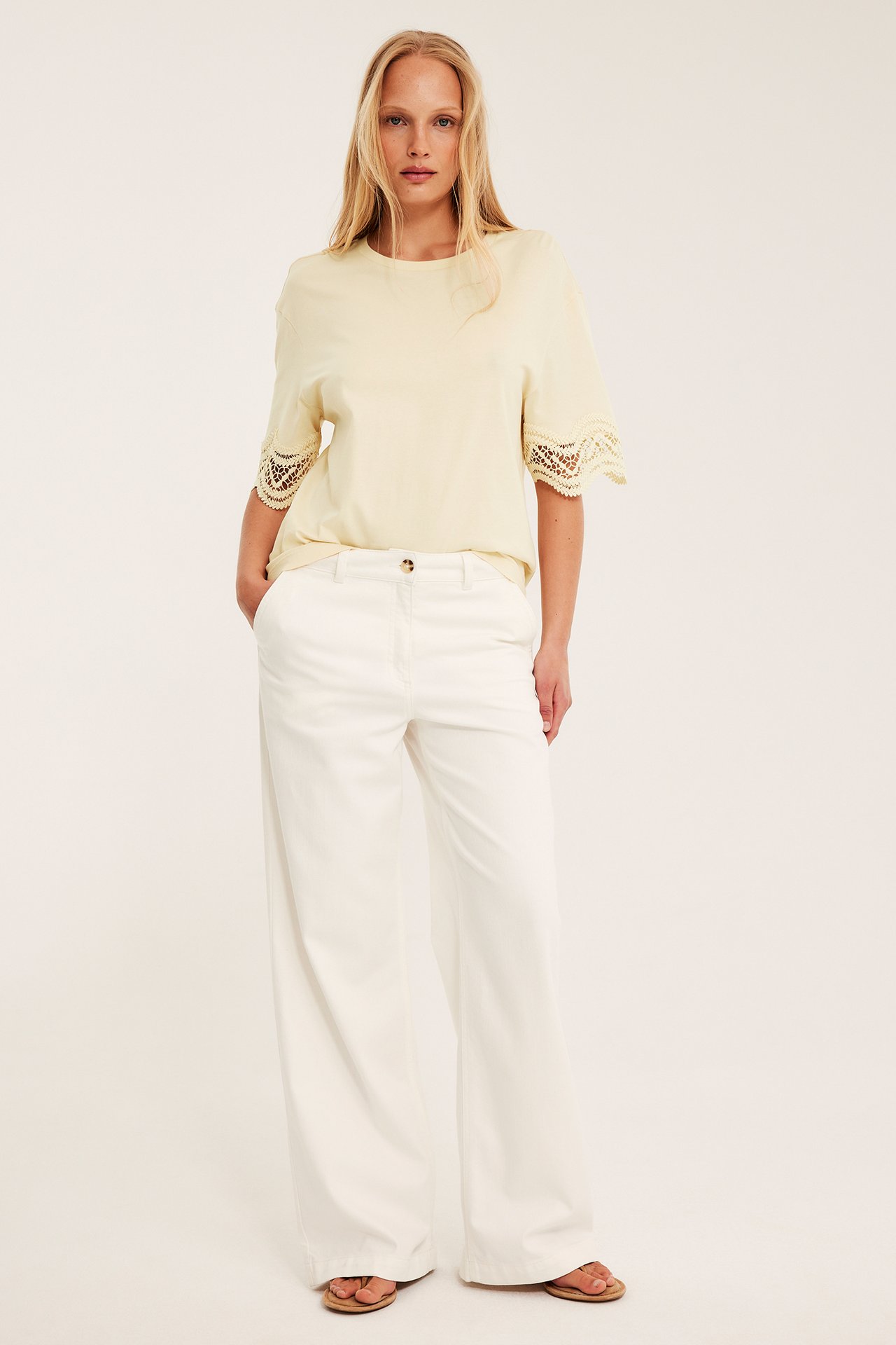 Framifrån: Kvinna i lös ljusgul t-shirt med spetsärmar, vita vida byxor och sandaler.