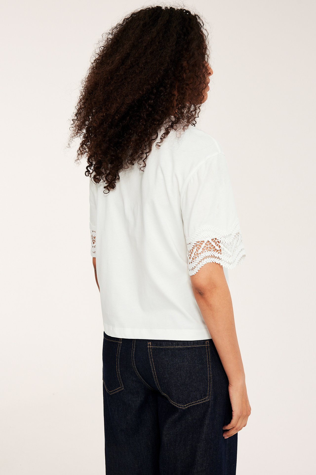 Naisen takasivuprofiili; löysä luonnonvalkoinen t-paita pitsihihoilla ja tummansiniset farkut.