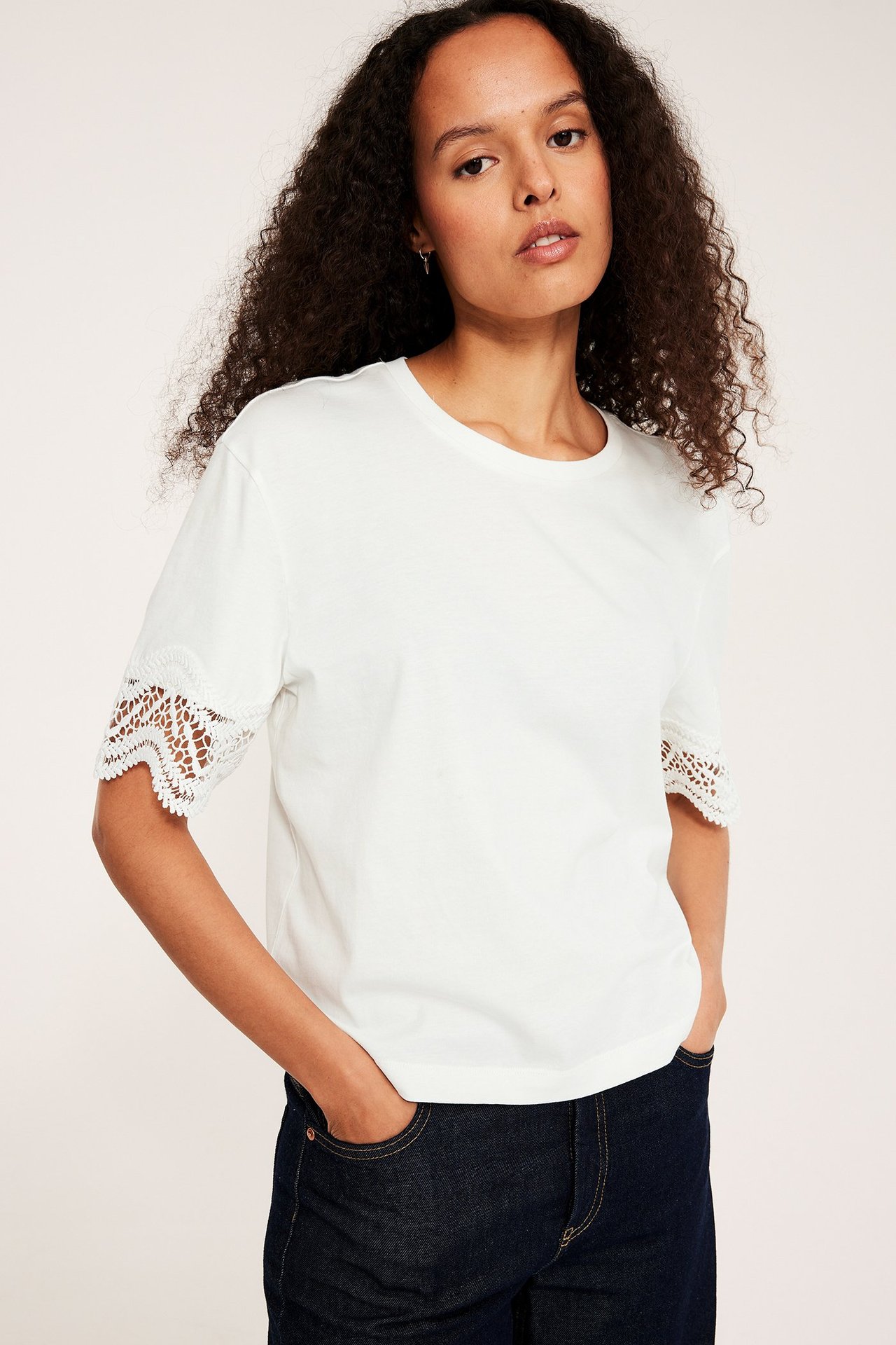 Edestäpäin, nainen yllään väljä offwhite t-paita pitsihihoilla ja tummansiniset farkut.