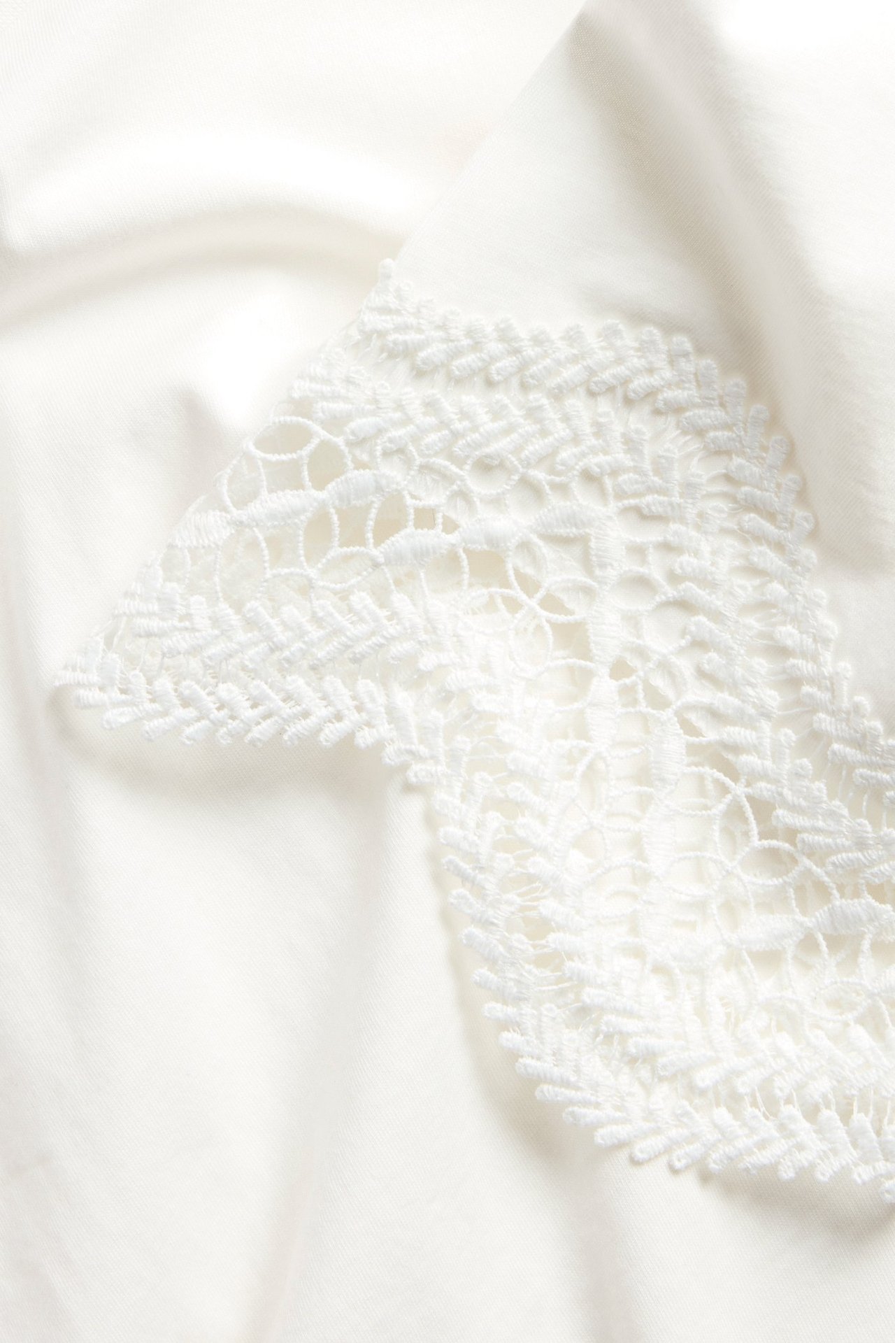 Zbliżenie na kremowy T-shirt, podkreślające jego misterny biały koronkowy brzeg.