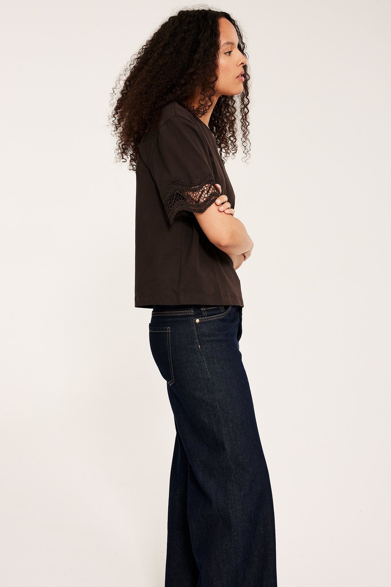 Sivukuva naisesta yllään löysä tummanruskea t-paita pitsihihoilla ja tummansiniset farkut.