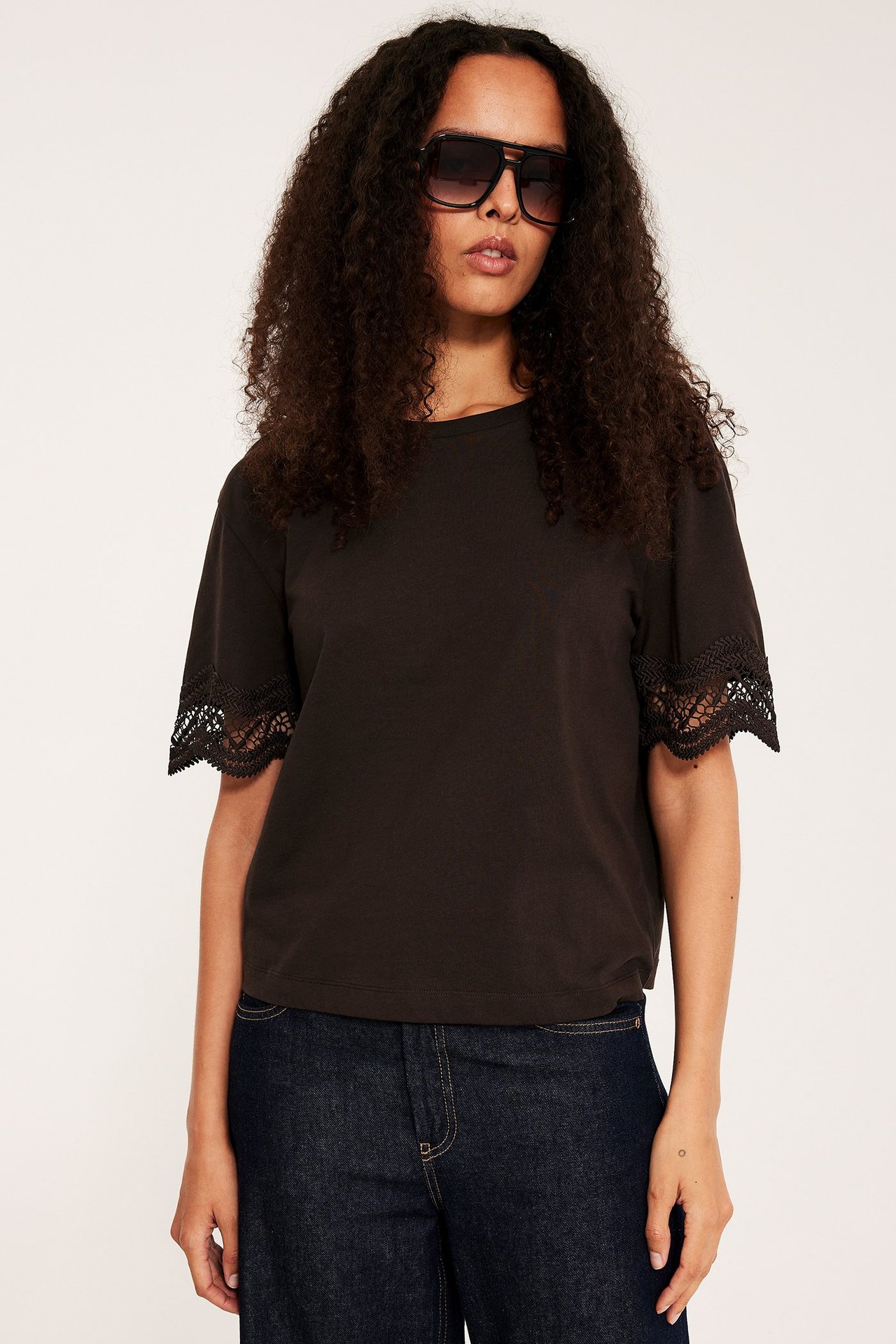 Edestä, nainen yllään tummanruskea löysä t-paita pitsihihoilla, tummat farkut ja mustat aurinkolasit.