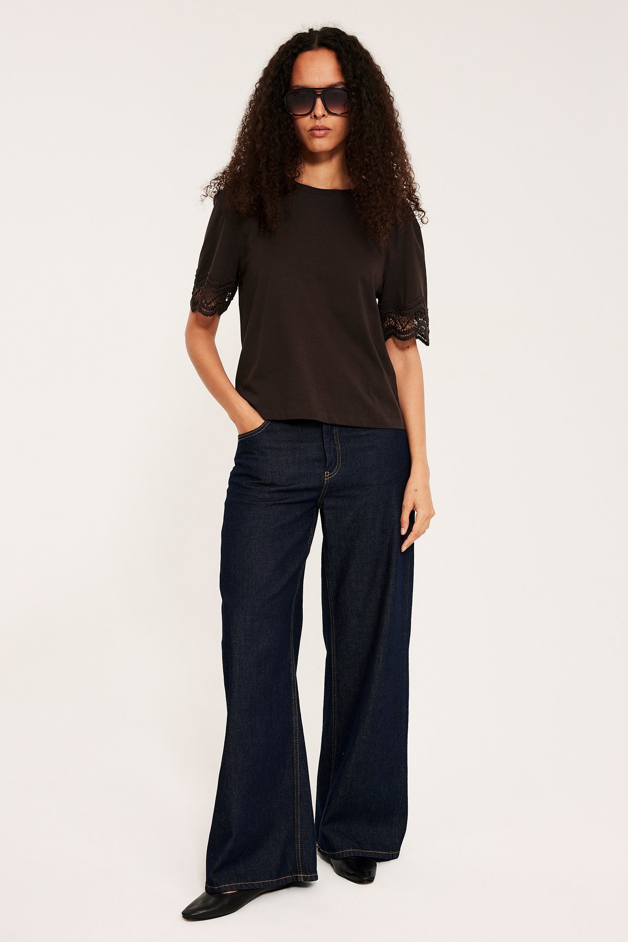 Nainen edestäpäin yllään löysä tummanruskea t-paita pitsihihoilla, tummansiniset flared-farkut ja aurinkolasit.