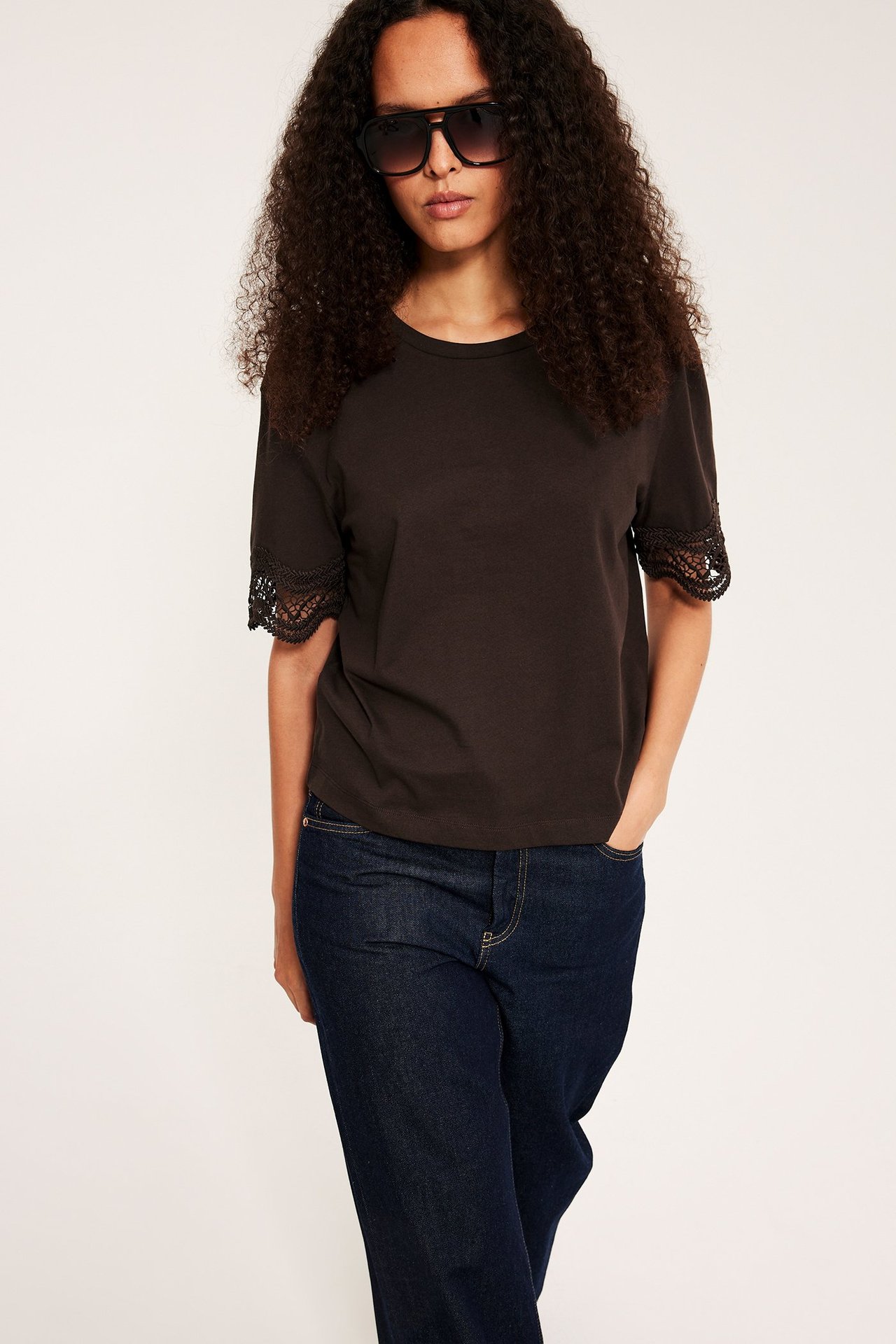 Edestäpäin nainen yllään tummanruskea löysä t-paita pitsihihoilla, tummansiniset farkut ja mustat aurinkolasit.