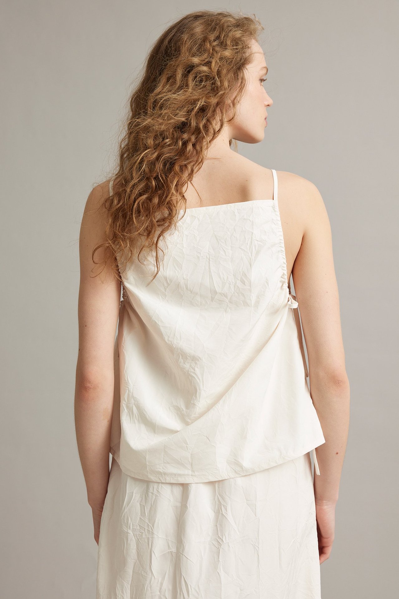 Bakfra: kvinne i hvit, teksturert polyestertanktopp med justerbare stropper og drapdetaljer.