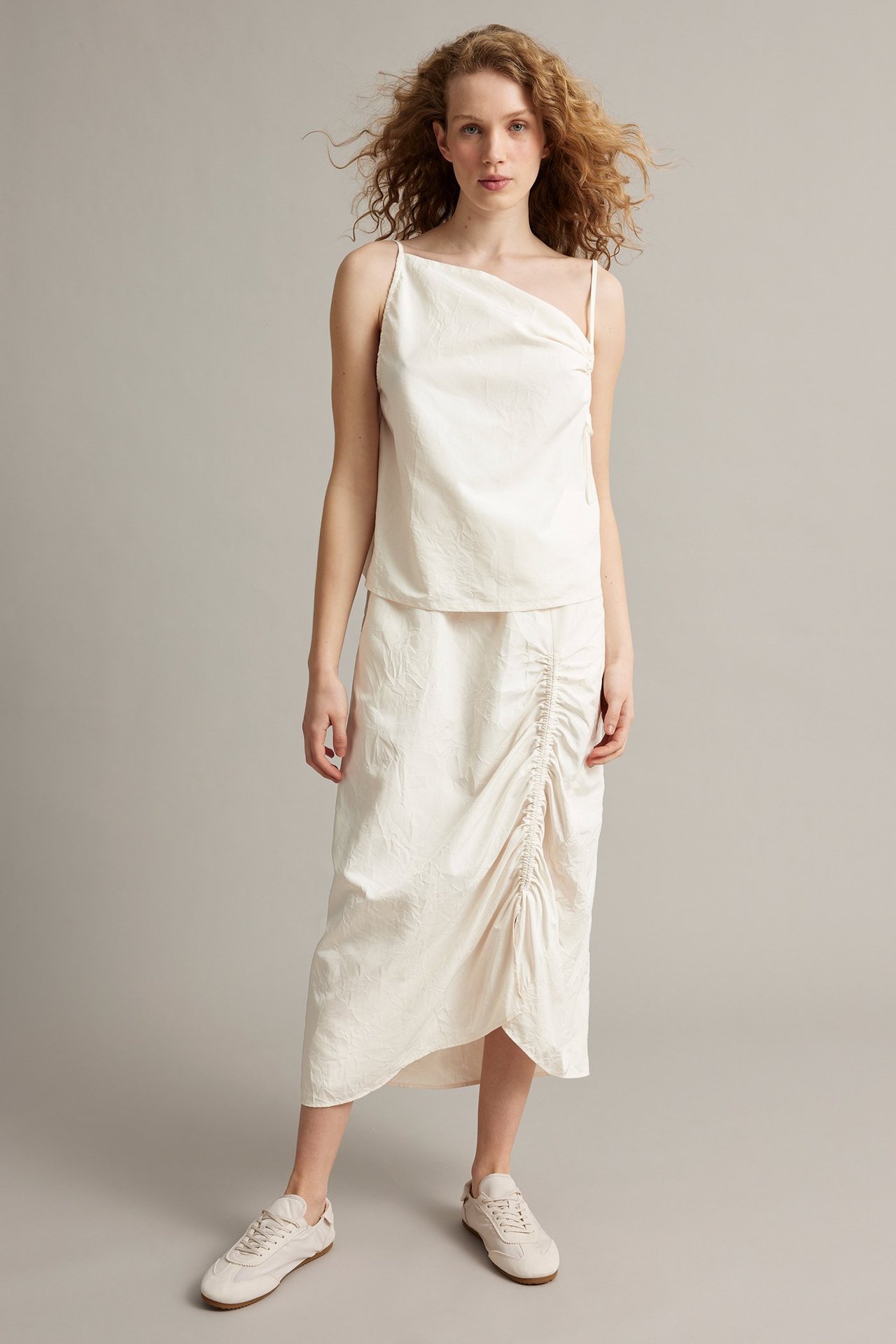 Forfra: kvinne i hvit drapert singlet, hvitt rynket midiskjørt og hvite sneakers.