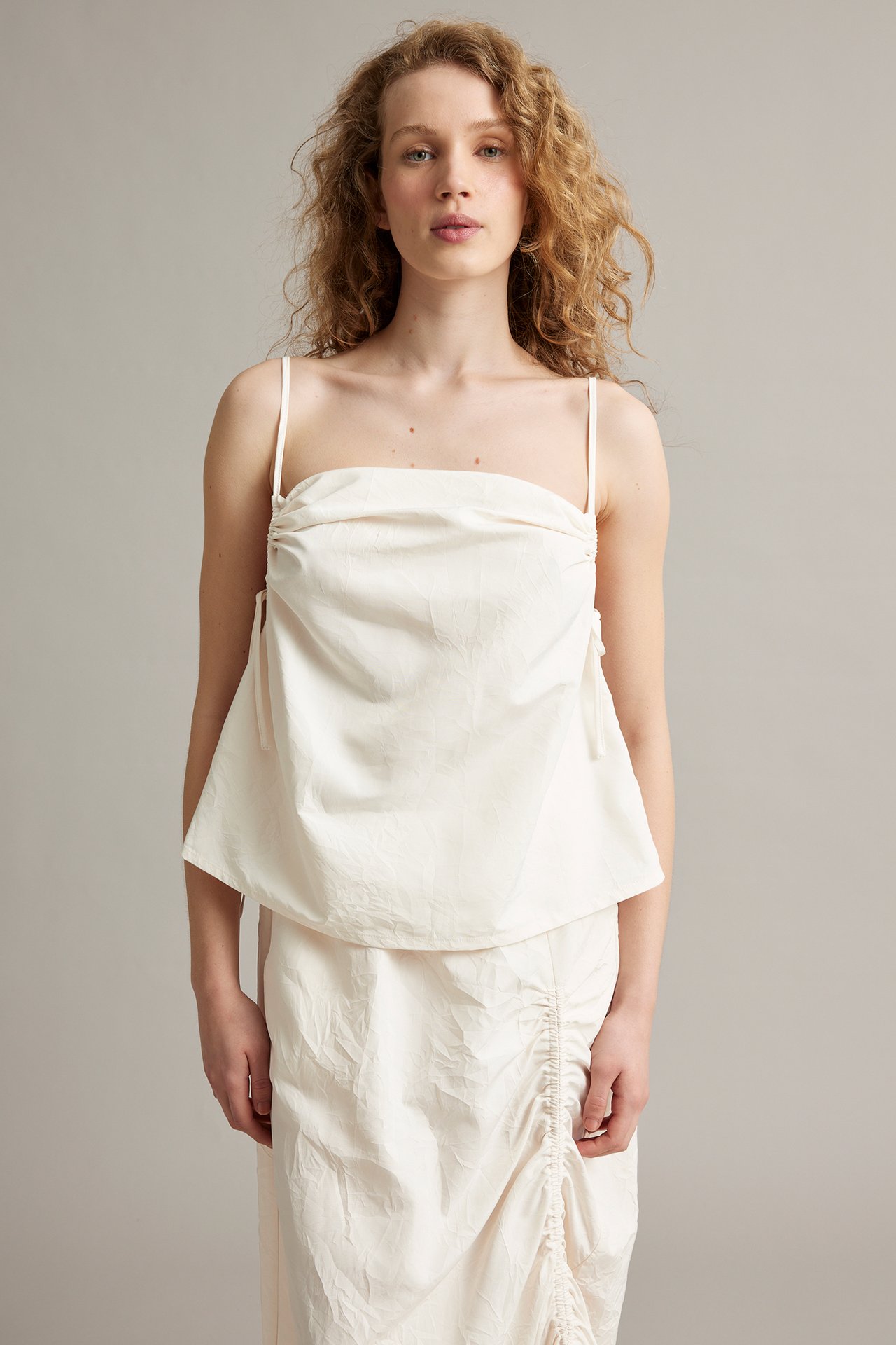 Forfra en kvinne i hvit drapert camisole-topp med spaghettistropper og et matchende rynket midiskjørt.