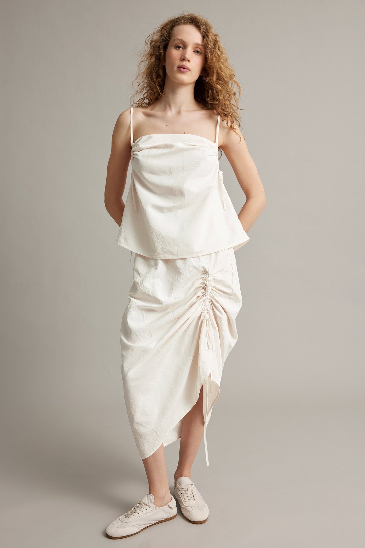 Framifrån, kvinna i vit draperad kjol med sidkanal, vitt linne och vita sneakers.