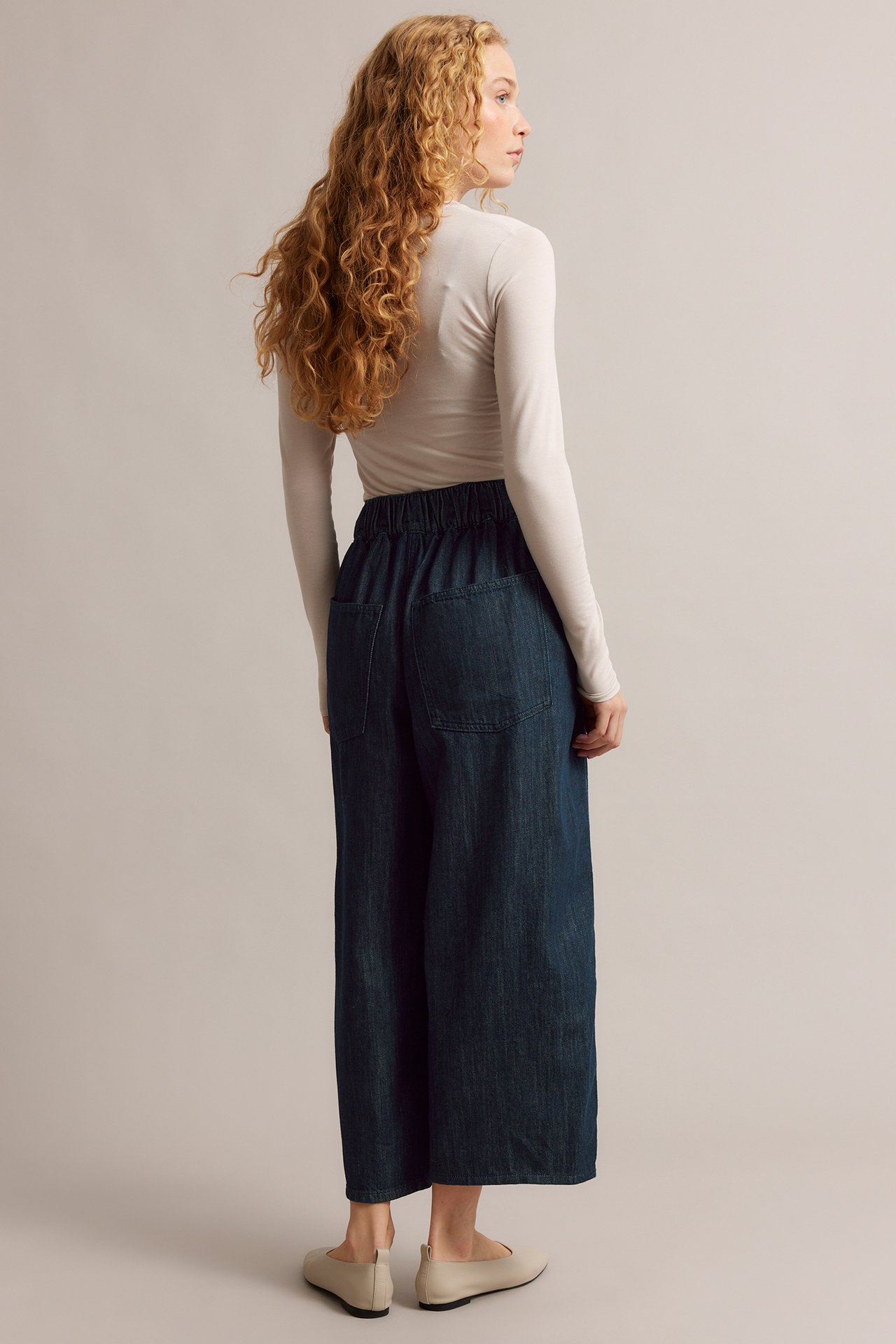 Kobieta od tyłu: ciemnoniebieskie jeansy barrel leg, beżowy top z długim rękawem i jasnobeżowe baleriny.